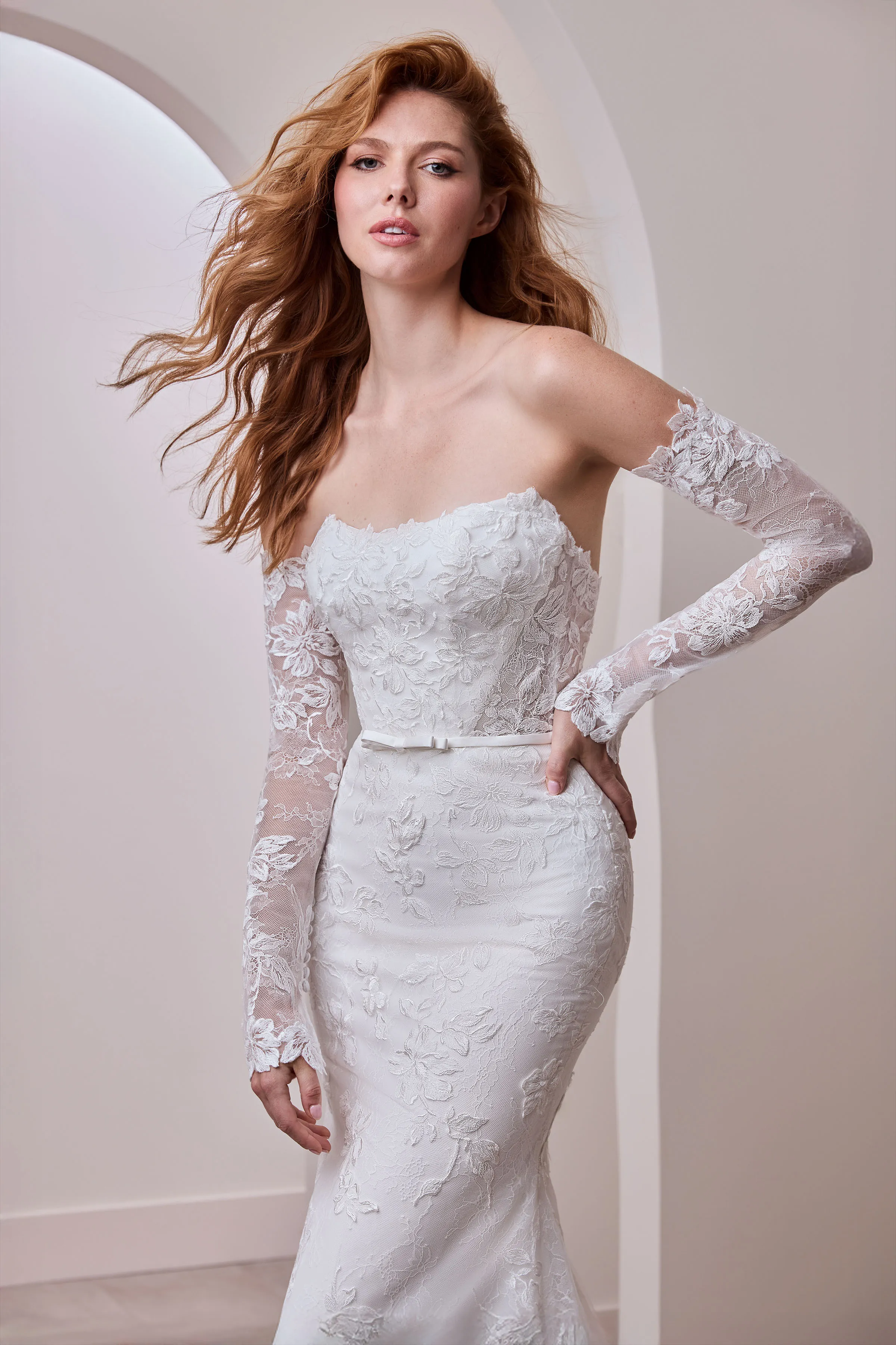 Close up view of a strapless lace wedding dress with a scoop neckline, separate long lace sleeves, a band with a bow at the waist, and a lace skirt, indoors in front of multiple arched walls.