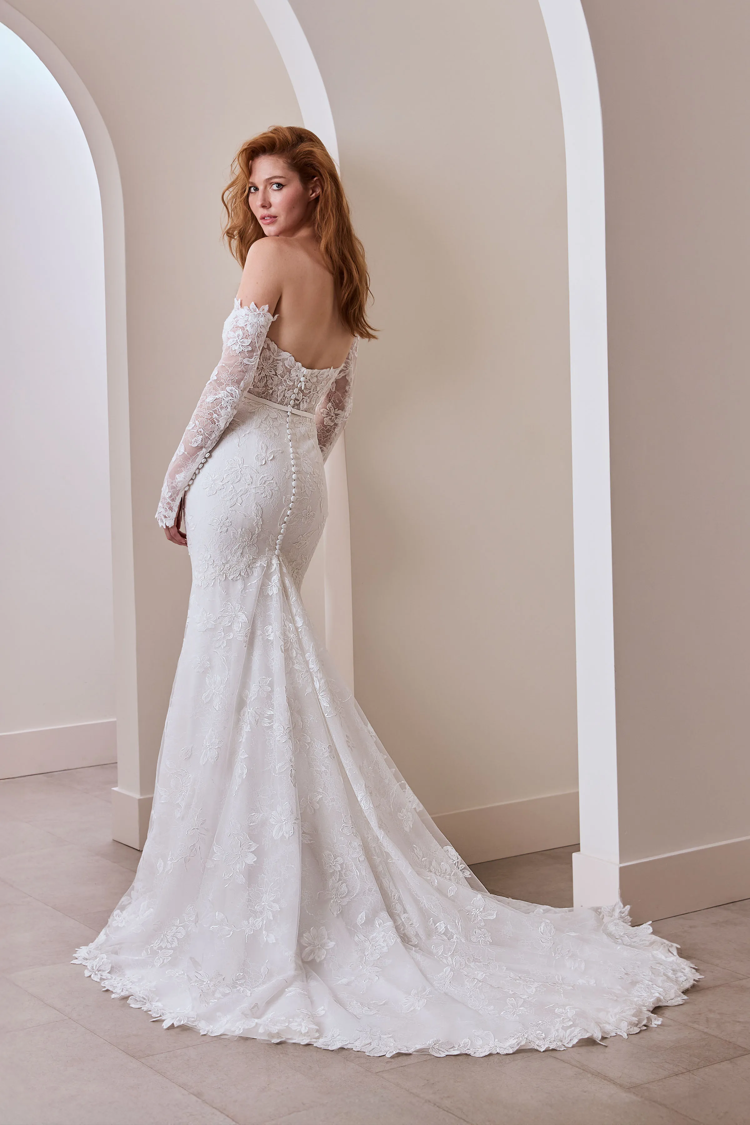 Back view of a strapless lace wedding dress with buttons partway down the back of the dress, with separate lace sleeves and a fit and flare lace skirt, indoors in front of multiple arched walls.