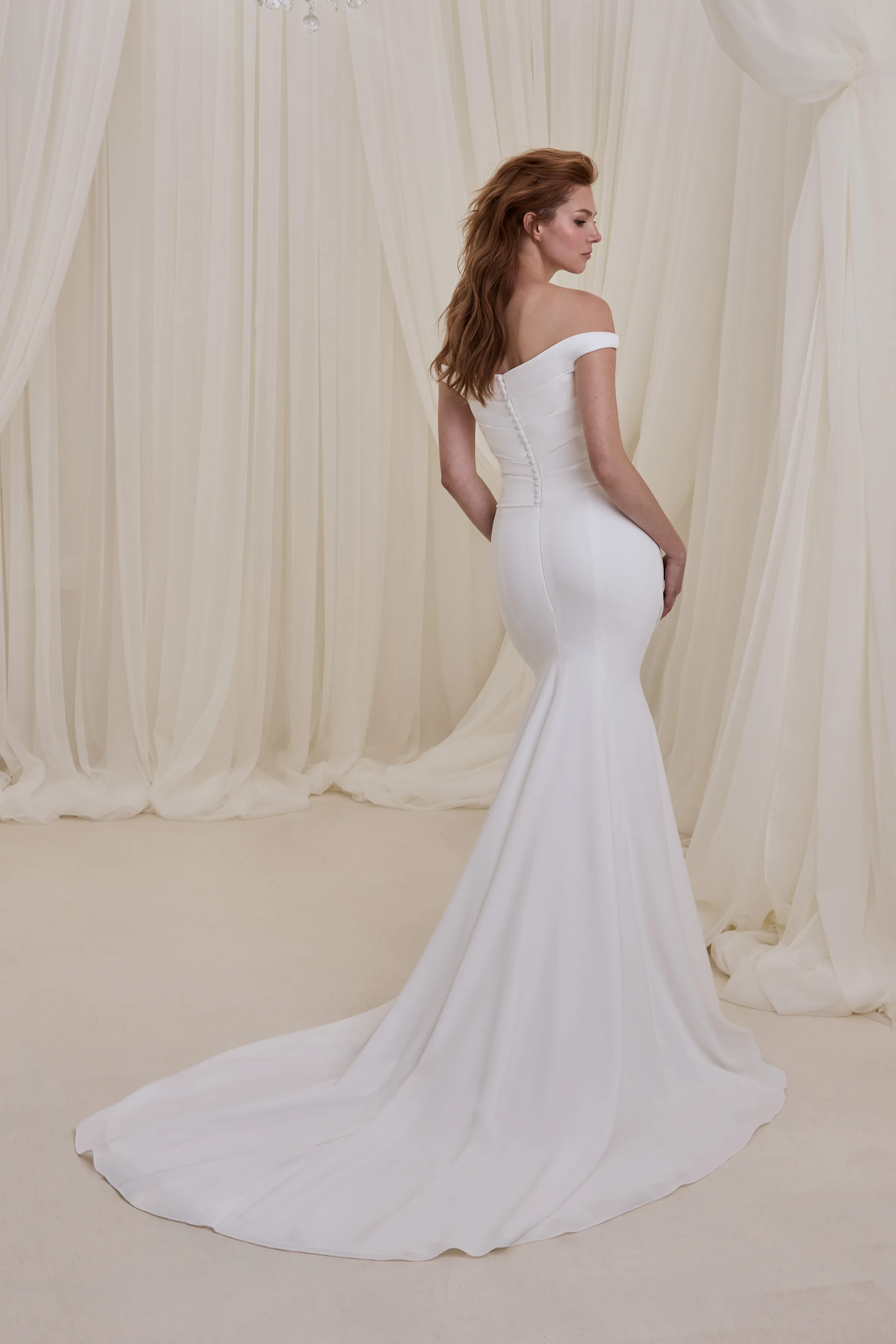 Back view of a fit and flare Crêpe wedding dress with an off the shoulder neckline and draped bodice, with buttons on the back of the bodice, in front of a wall of tulle curtains.