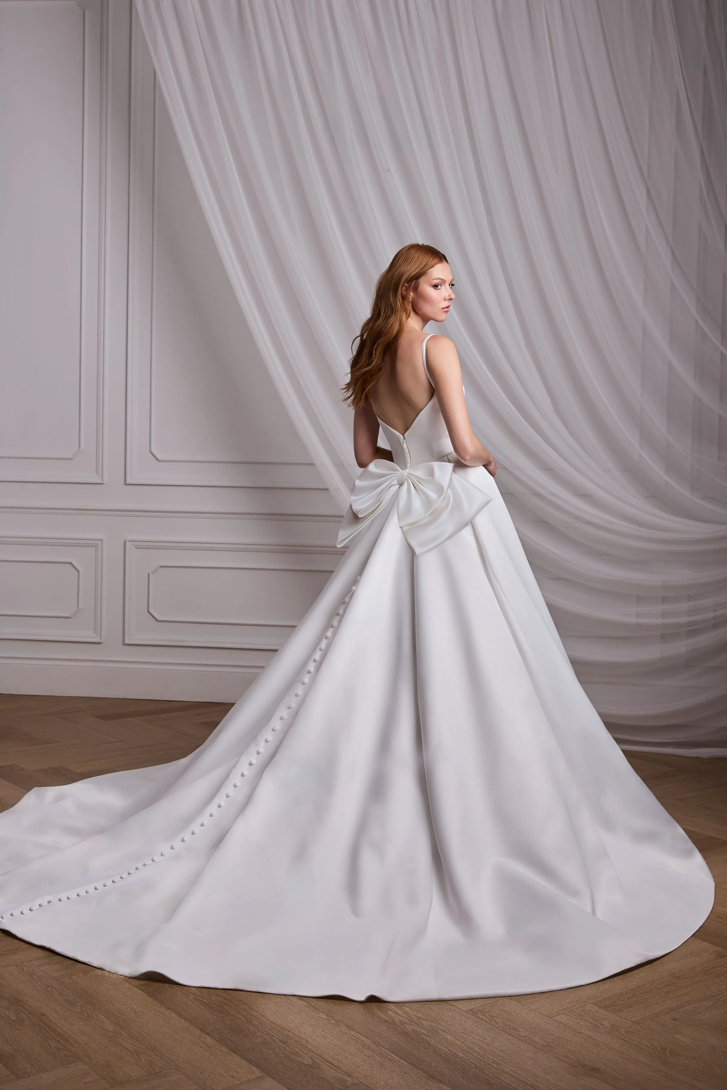 Back view of a sleeveless Satin Faille wedding dress with a back bow and buttons down the back of the dress, with a full circle skirt, in a ballroom with draped tulle in the background.