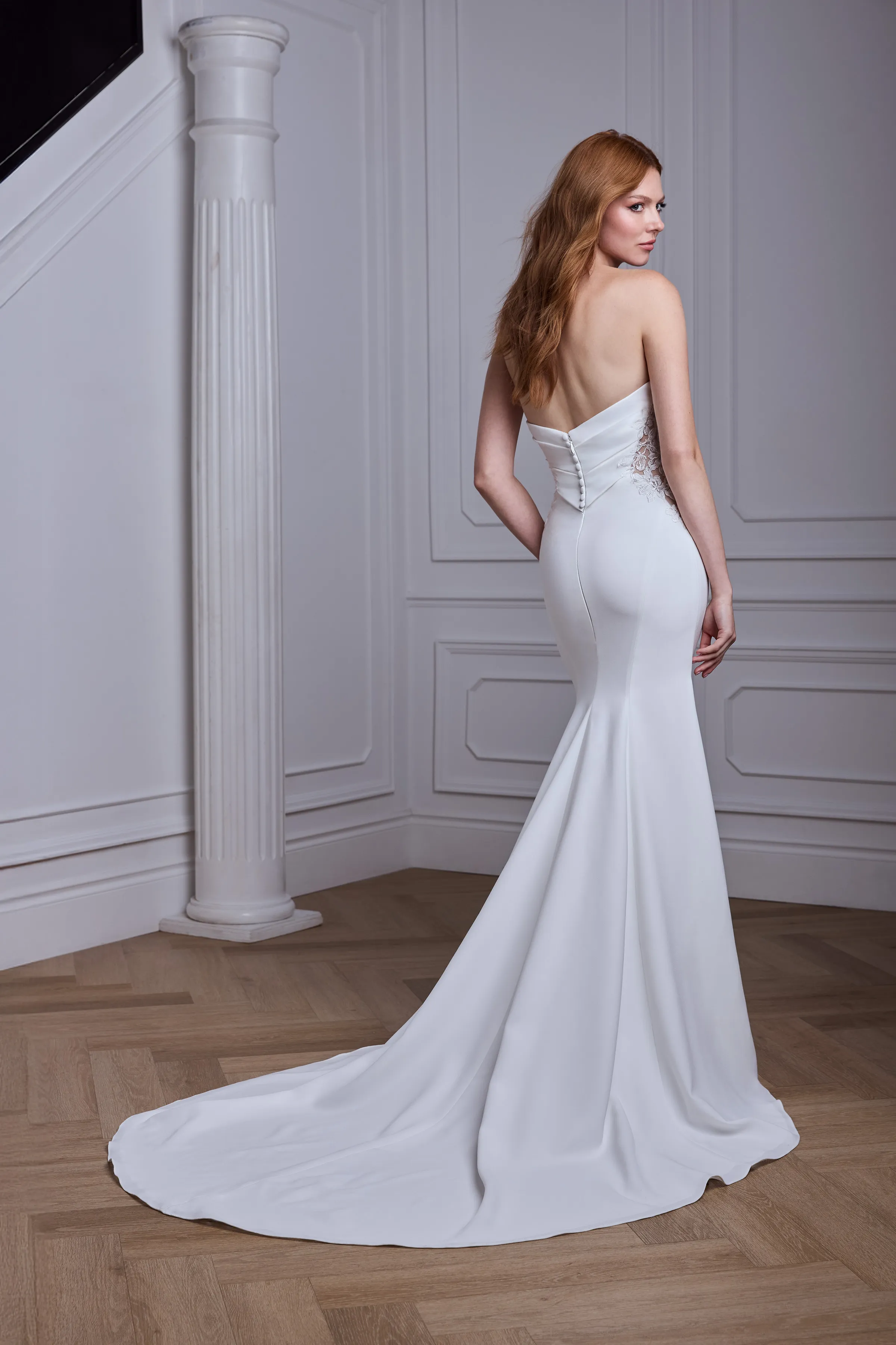 Back view of a strapless Crêpe wedding dress with a draped bodice, lace cut-outs on the side seams, and buttons down the back of the bodice, with a fit and flare skirt, in a ballroom with a pillar in the background.