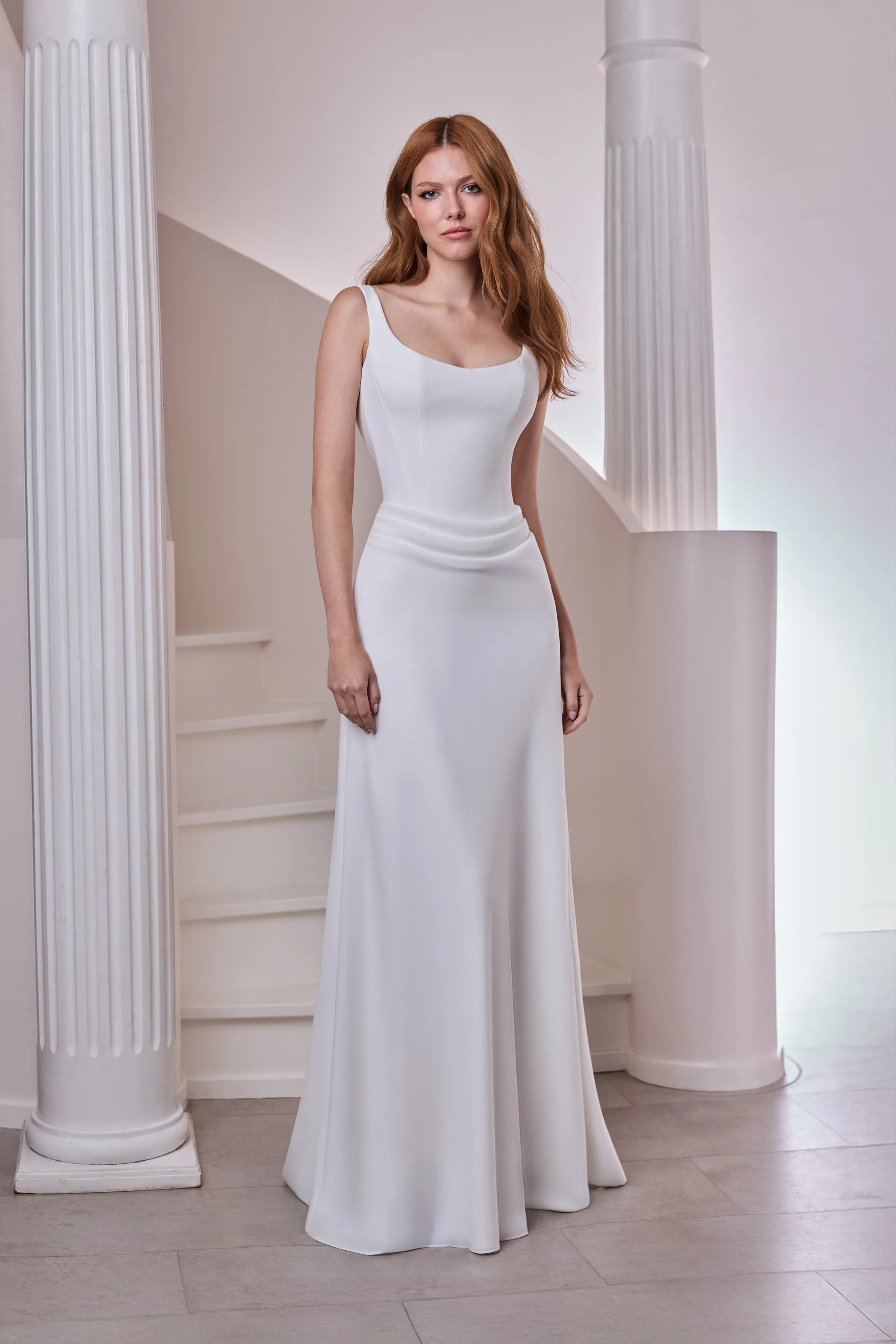 Front view of a sleeveless drop waisted Crêpe wedding dress with a scoop neckline, seamed bodice, and a draped A-line skirt, indoors in front of a spiral staircase and pillars.