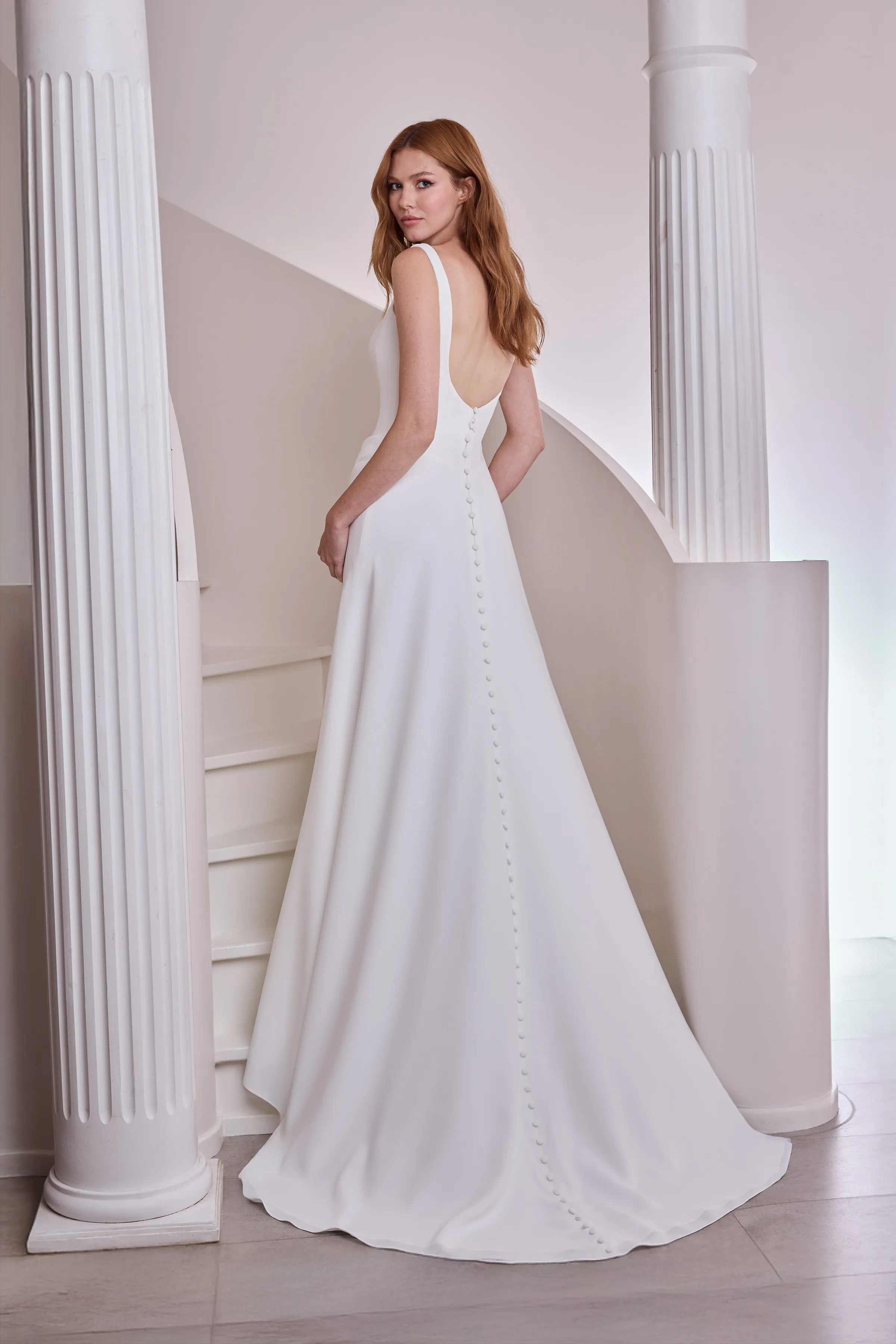Back view of a sleeveless drop waisted Crêpe wedding dress with buttons down the back of the dress, and an A-line skirt, indoors in front of a spiral staircase and pillars.