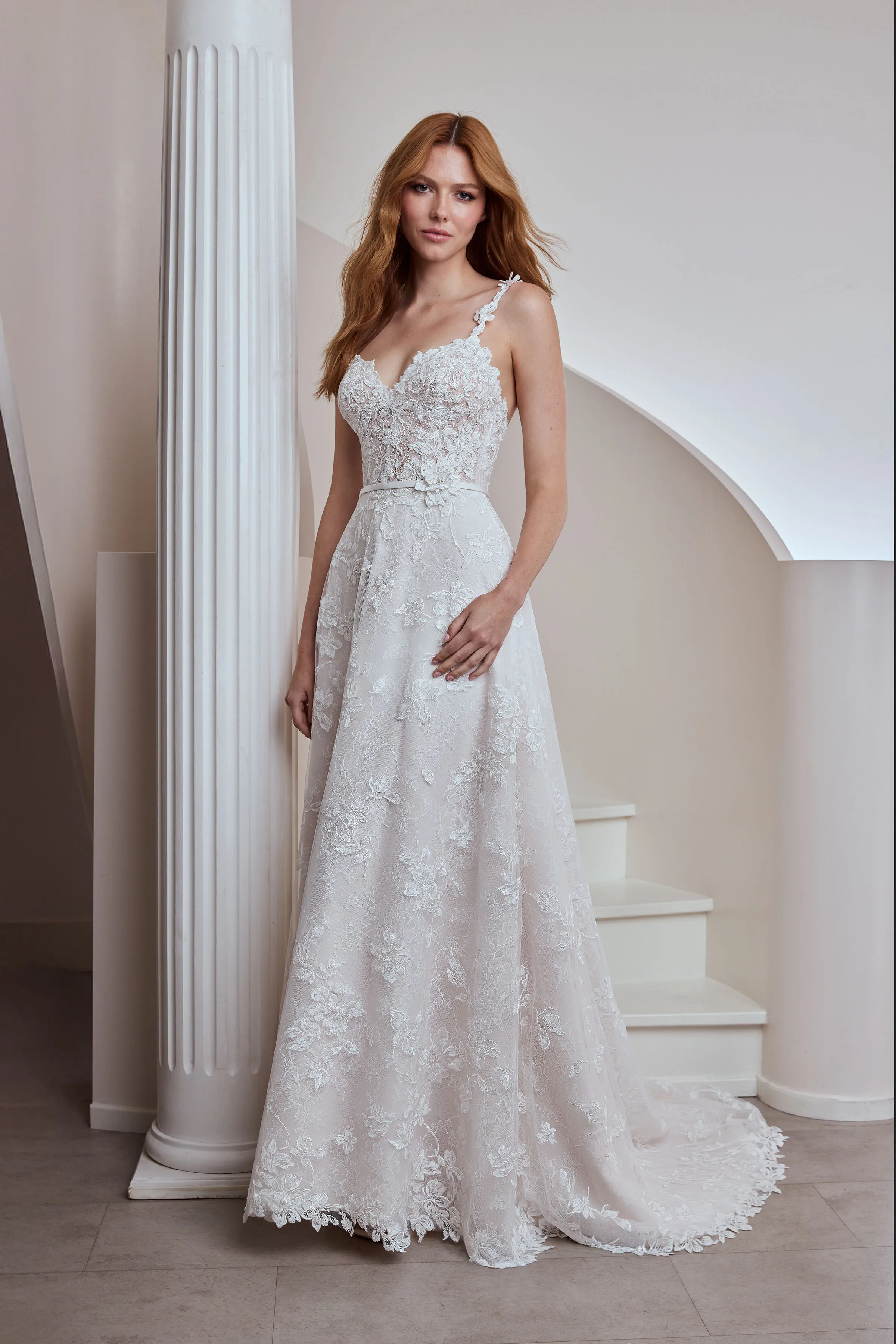 Front view of a Lace wedding dress with lace straps and a sheer bodice, with a floral embellishment waist band, and an A-line skirt, indoors in front of a spiral staircase and a pillar. 