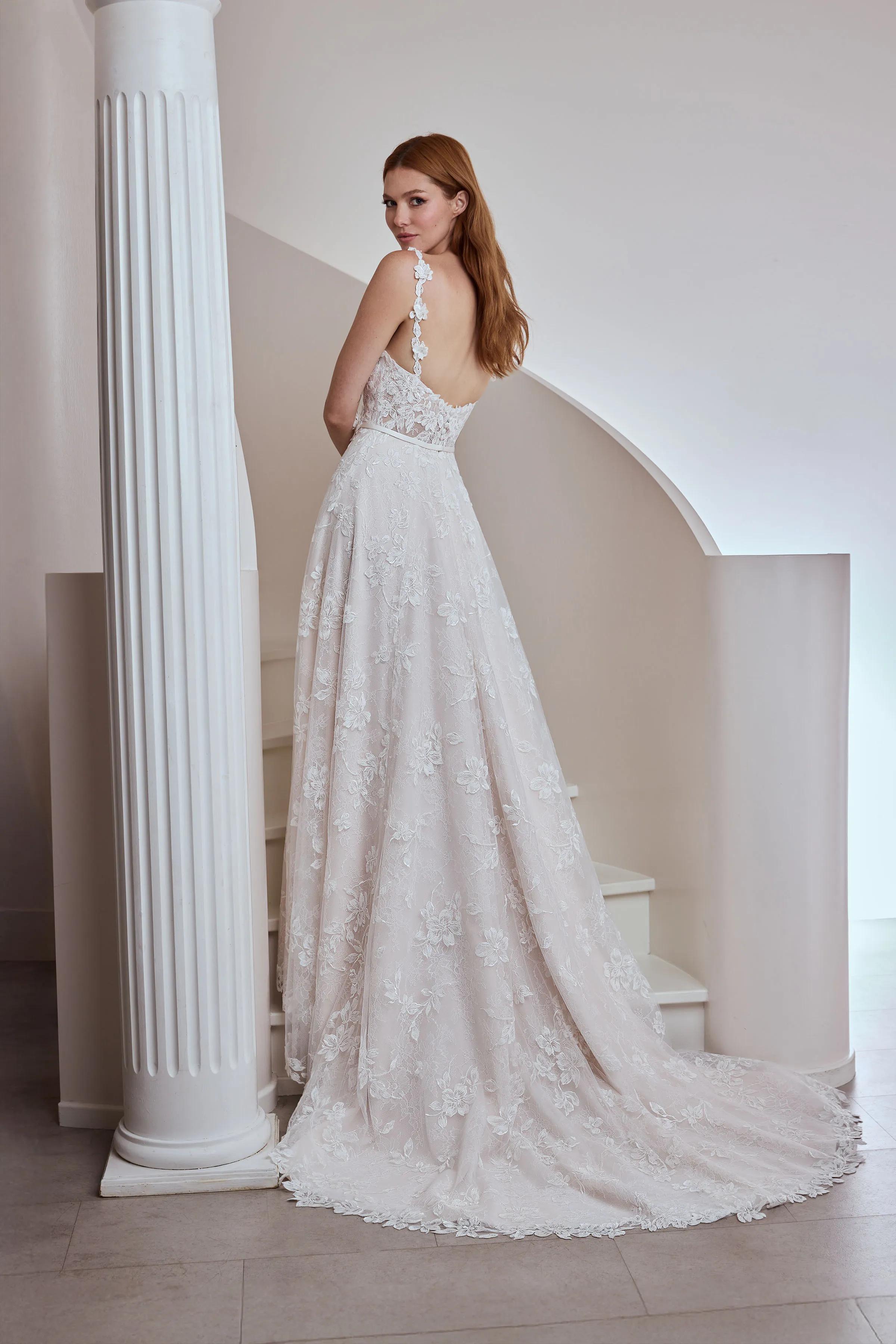 Back view of a Lace wedding dress with lace straps and a sheer bodice, with buttons on the back of the bodice and a band at the waist, with an A-line skirt, indoors in front of a staircase and a pillar.