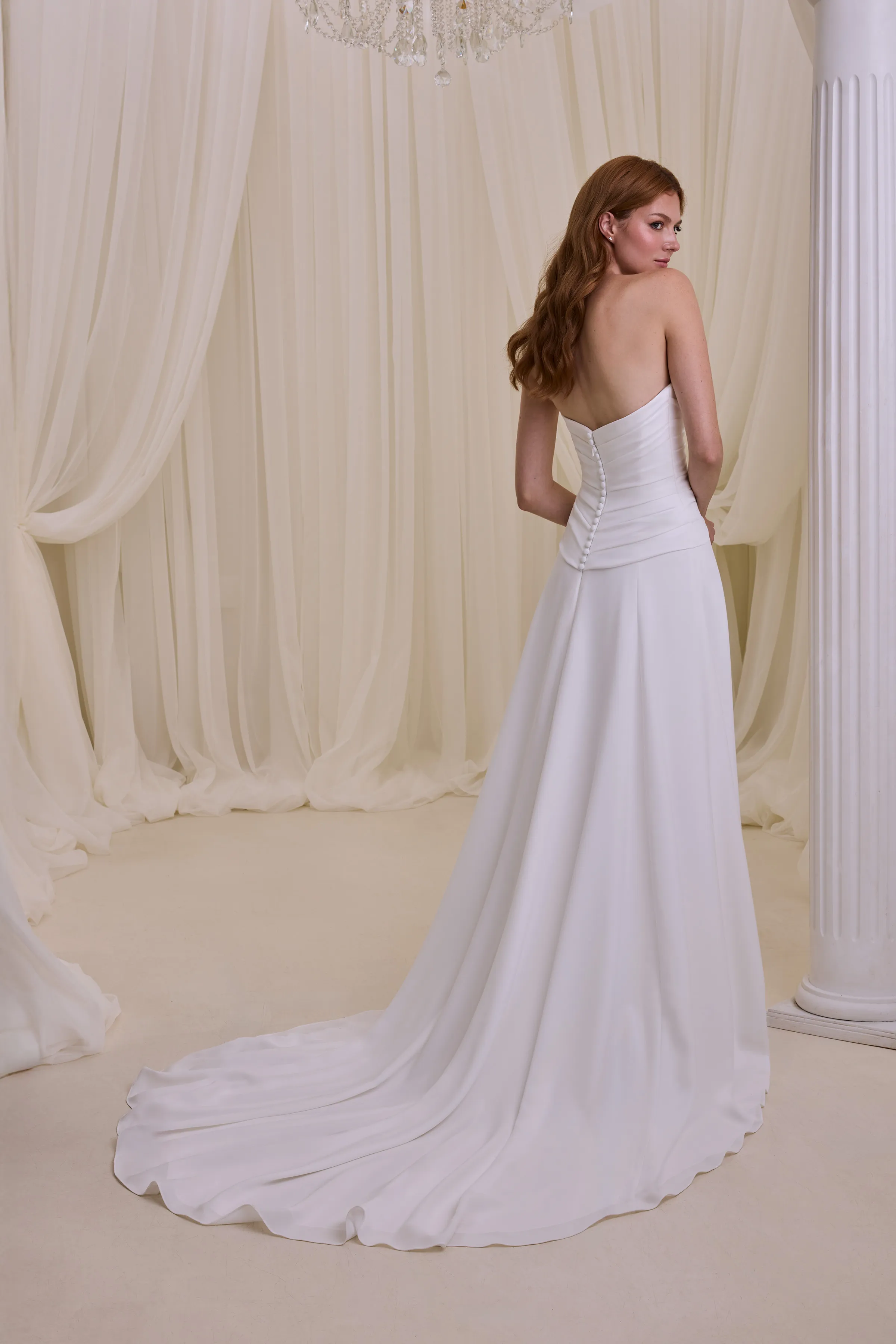 Back view of a strapless Crêpe wedding dress with a draped drop waisted bodice, buttons down the back of the bodice, and an A-line skirt, in front of a pillar and a wall of tulle curtains.