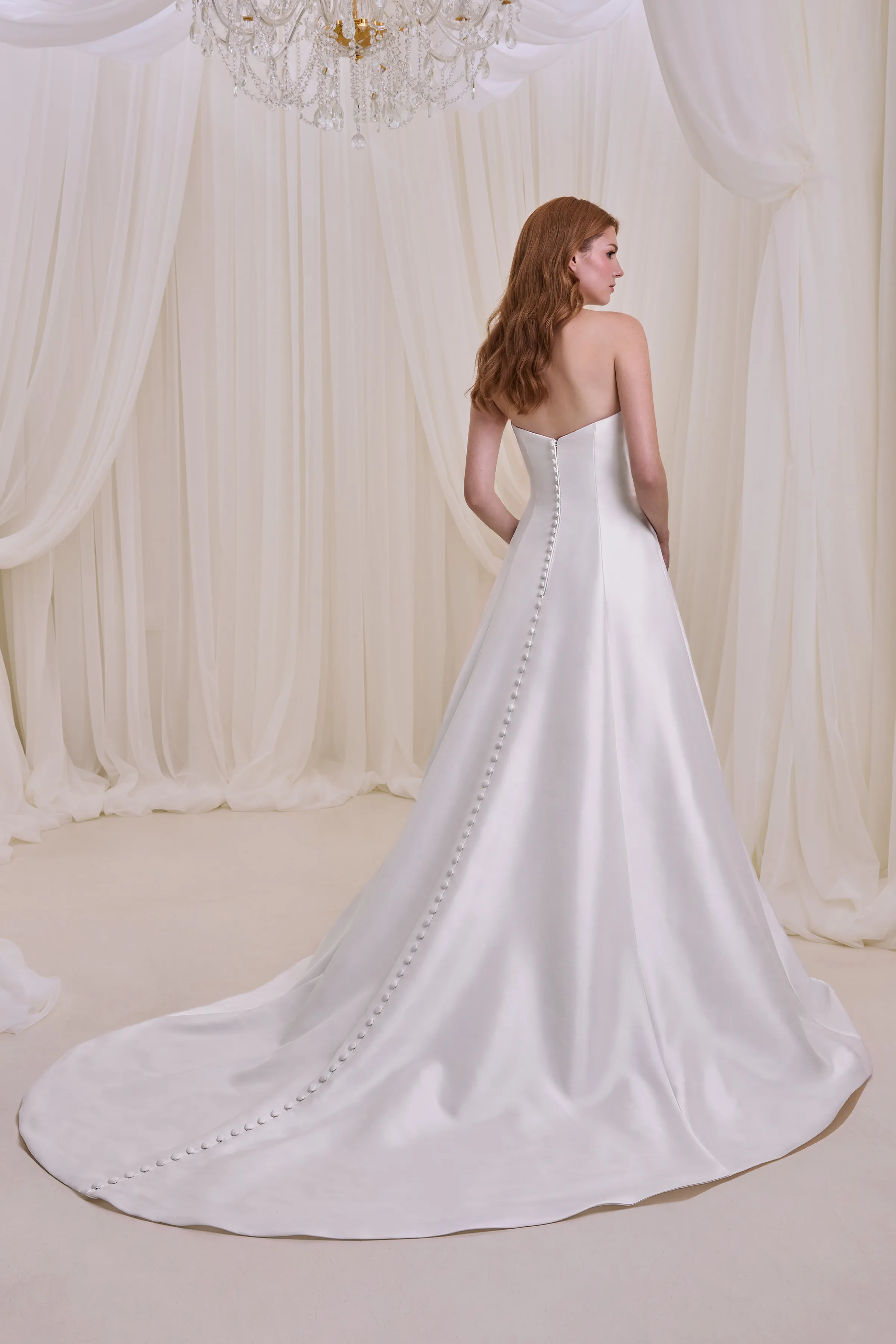 Back view of a strapless Satin wedding dress with buttons down the back of the dress to the hem and an A-line skirt, indoors in front of a wall of tulle curtains.