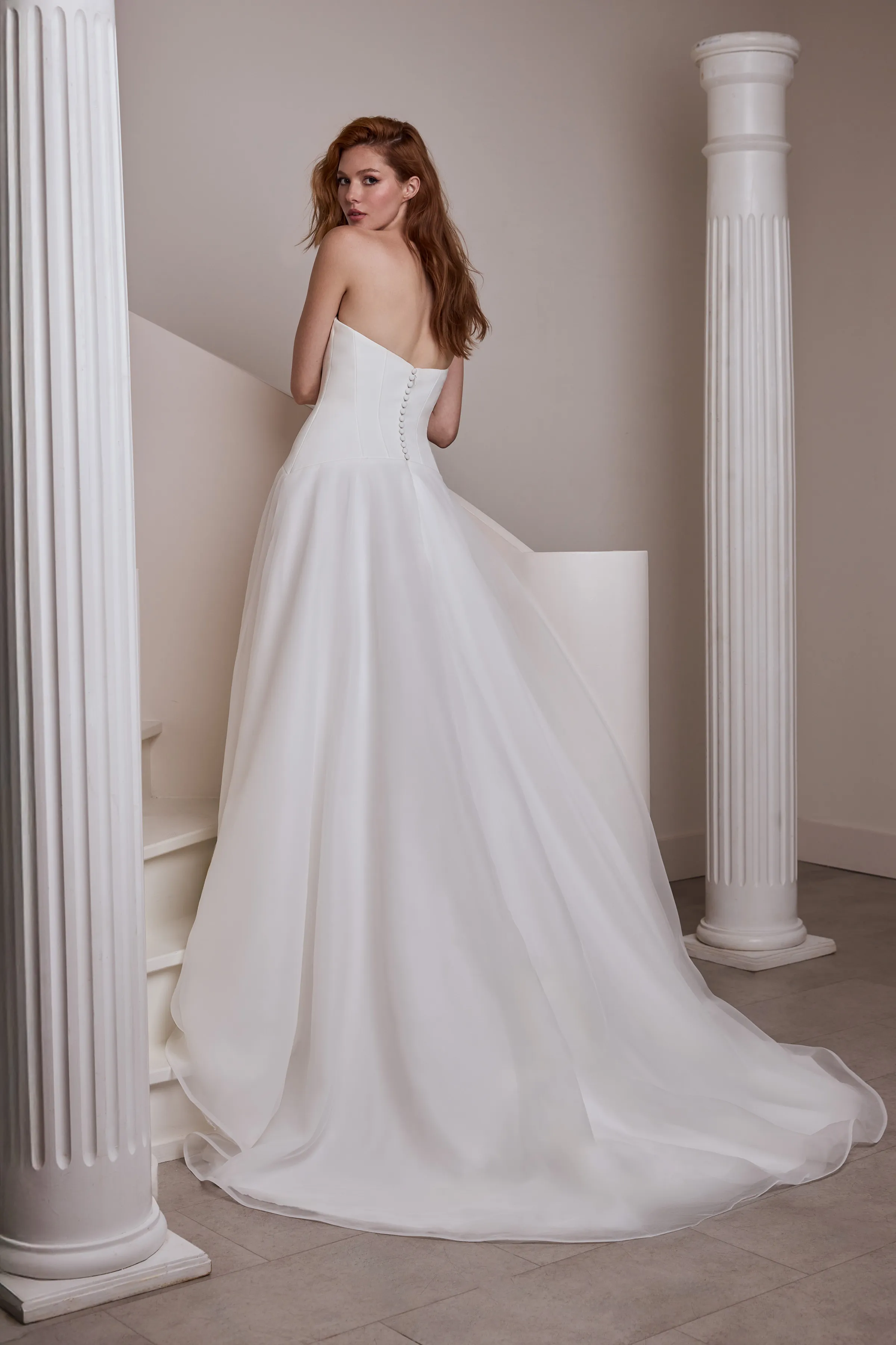 Back view of a strapless wedding dress with a drop waisted Crêpe seamed bodice, buttons down the back of the bodice, and curved pointed hem, with an Organza overskirt, in front of a spiral staircase and pillars.