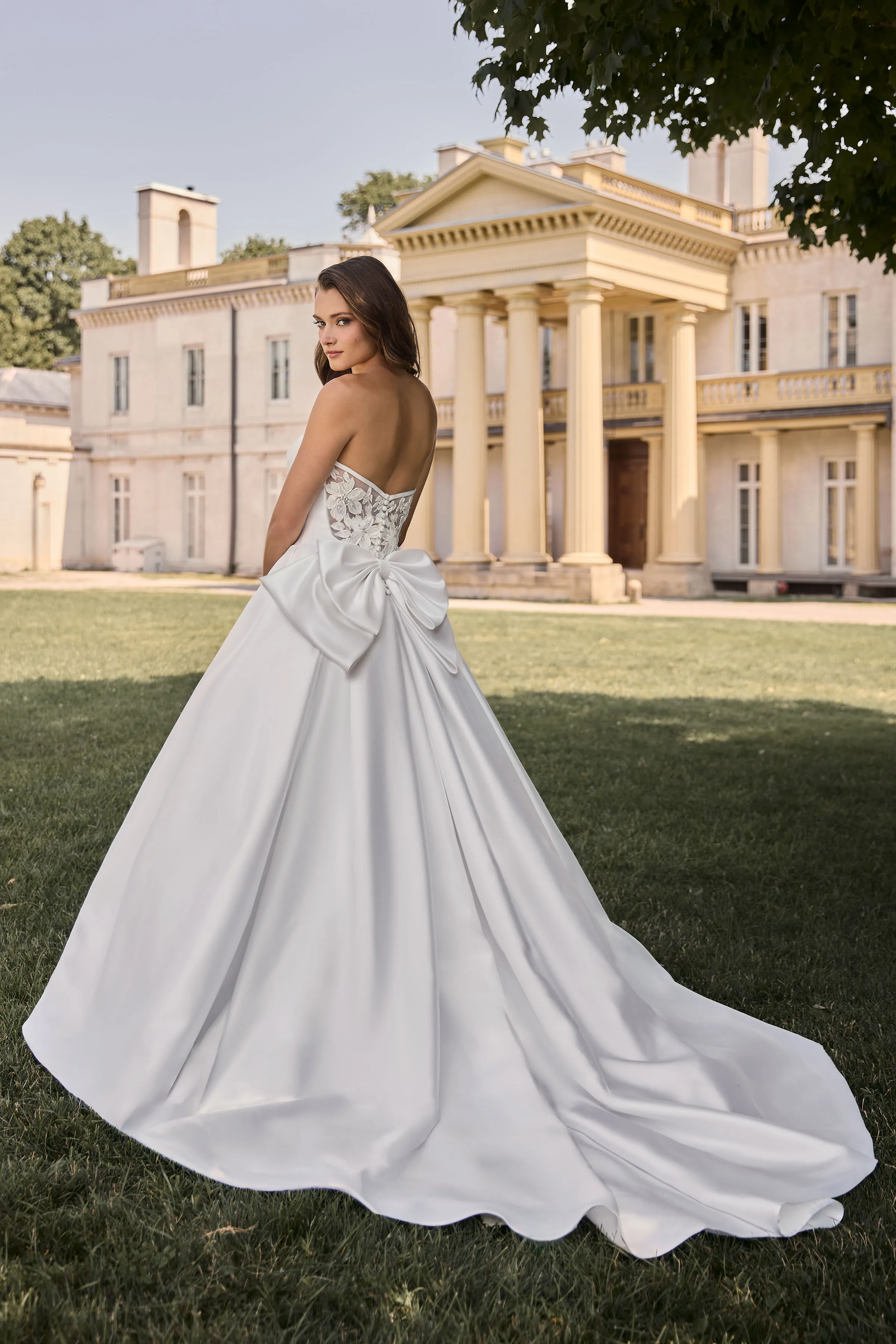 Back view of a strapless Satin ball gown wedding dress with a sheer lace back and a large double bow, outside in the grass in front of a large manor.
