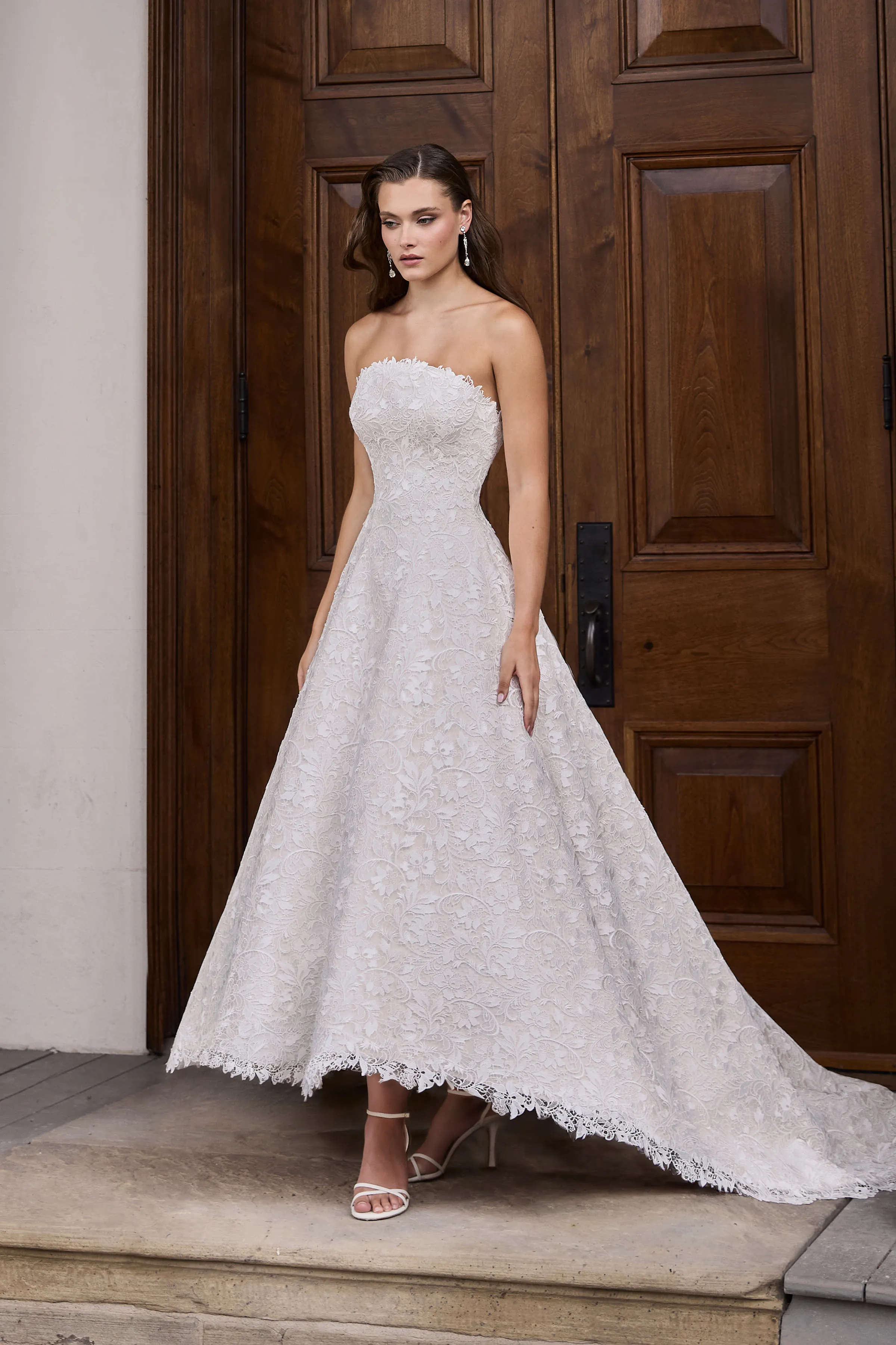 Front view of a strapless high-low Lace wedding dress with an arched neckline and A-line skirt, in front of a large wooden door.