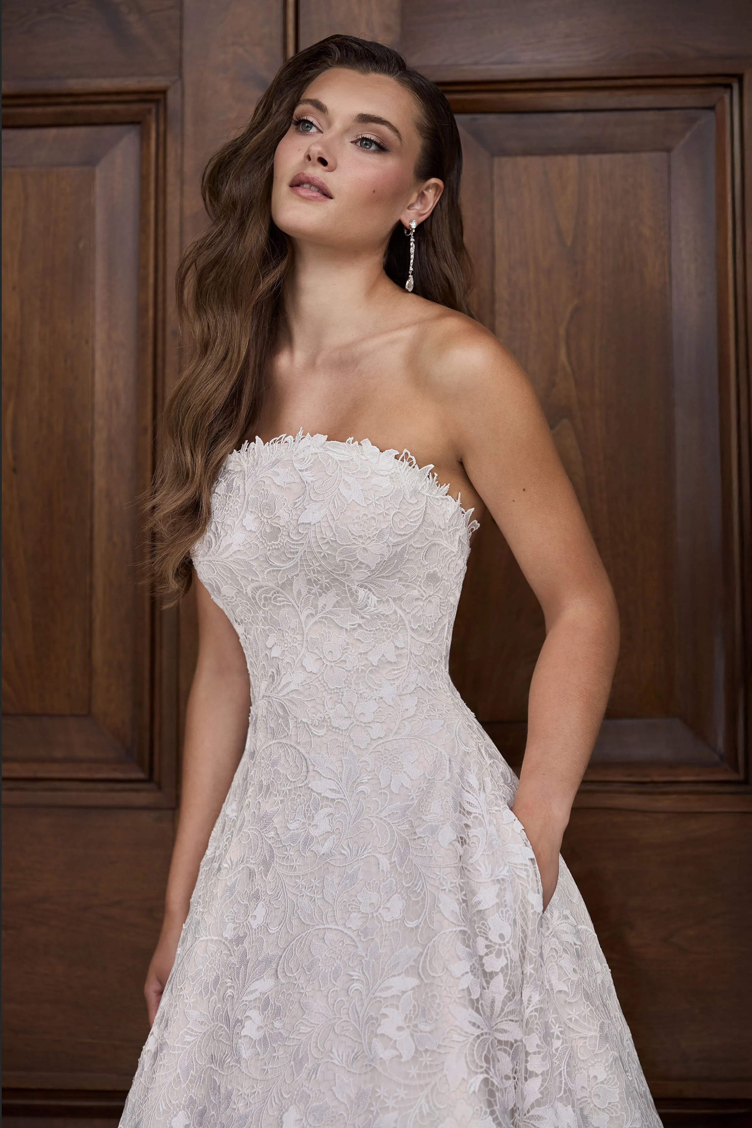 Close up view of a Lace wedding dress with an arched neckline and a lace skirt with pockets, in front of a large wooden door.