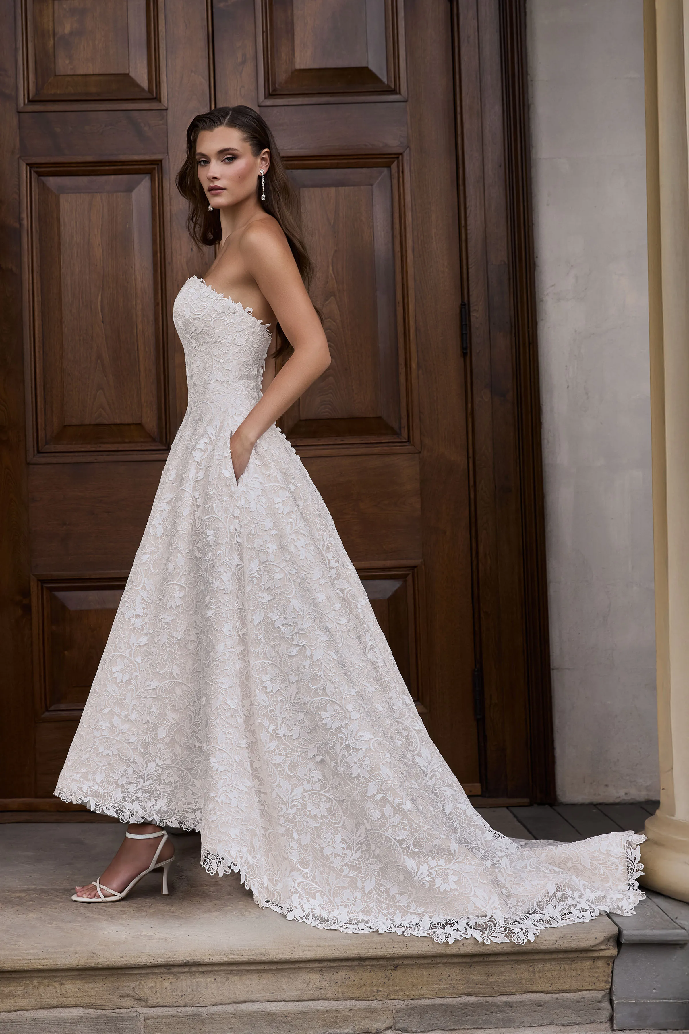 Side view of a strapless high-low Lace wedding dress with an A-line skirt with pockets, in front of a large wooden door.