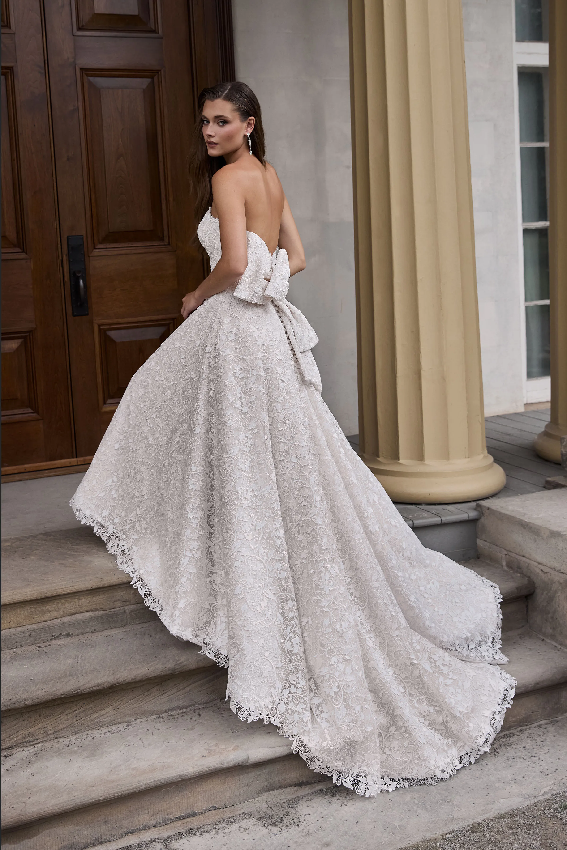 Back view of a strapless Lace wedding dress with an A-line skirt and large lace bow, on the steps of a building in front of a large wooden door.