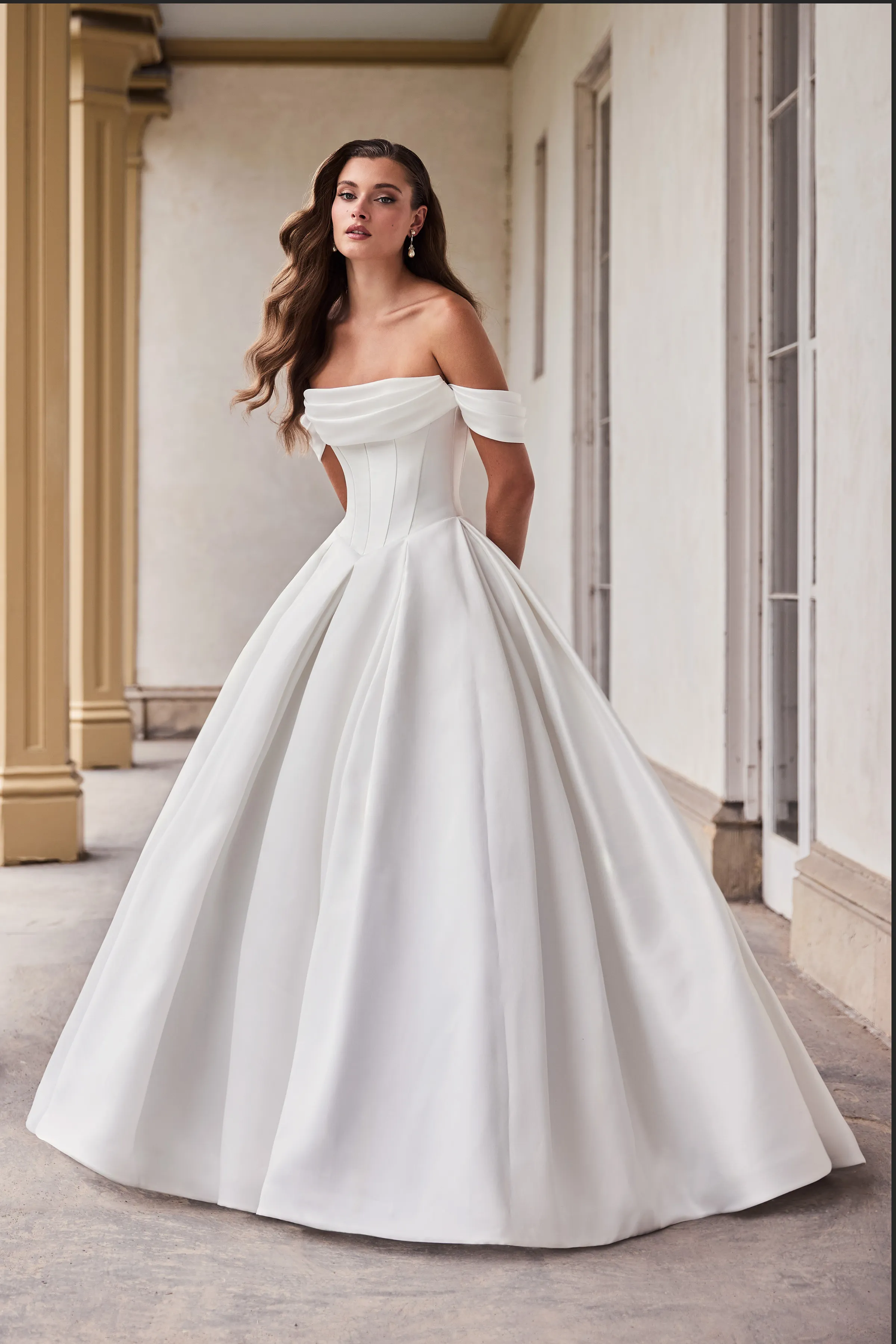 Front view of a strapless Mikado wedding dress with draped sleeves, a seamed bodice, a Basque waistline, and a pleated ball gown skirt, in front of pillars and a wall with windows.
