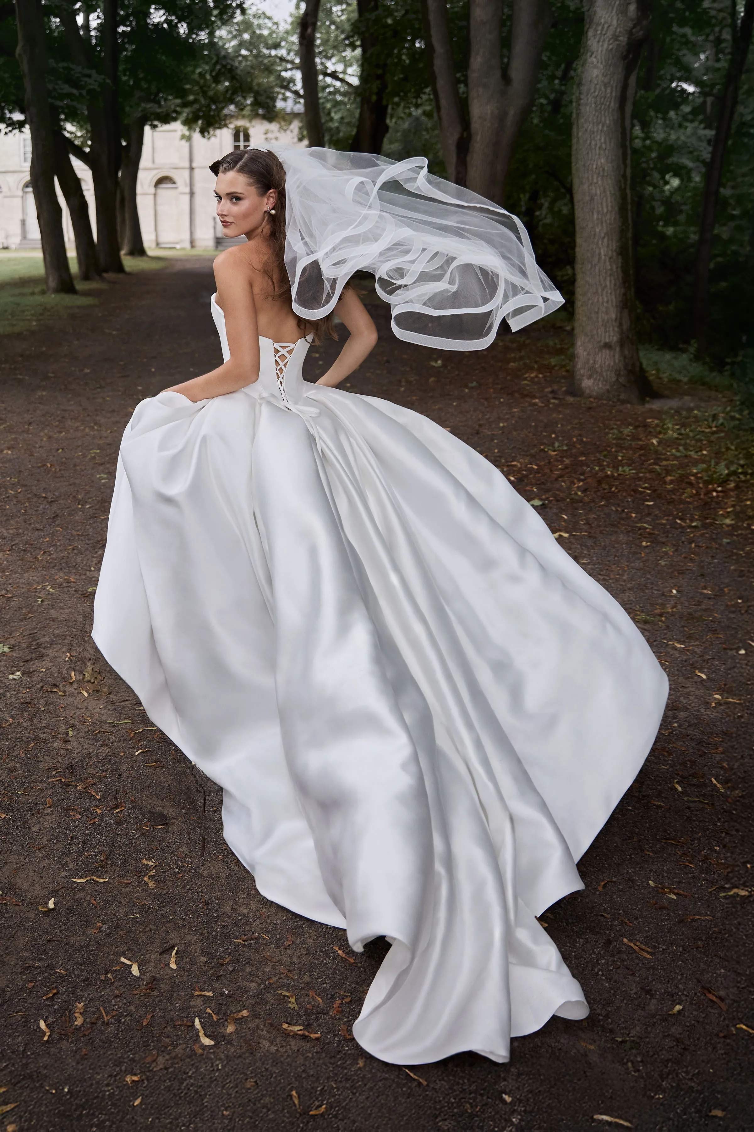 Back view of a strapless ball gown Mikado wedding dress with a lace-up back and a pleated skirt with pockets. The model is wearing a veil and running outside down a path lined with trees.