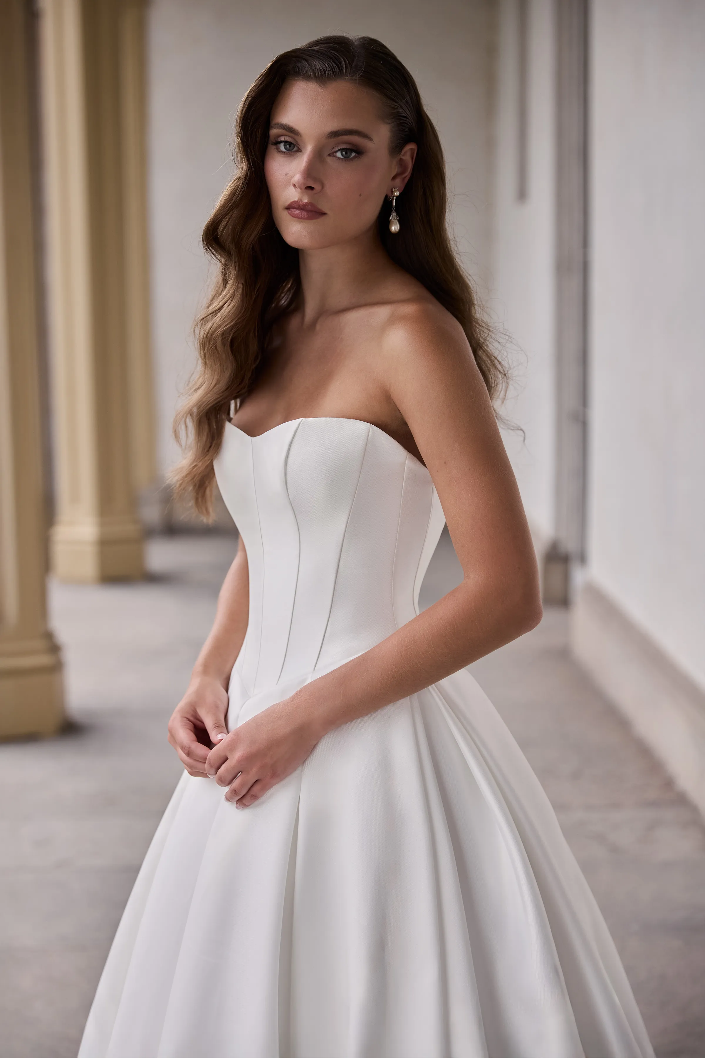 Close up view of a strapless Mikado wedding dress with a scoop neckline, a seamed bodice, a Basque waistline, and a pleated ball gown skirt, in front of pillars and a wall with windows.