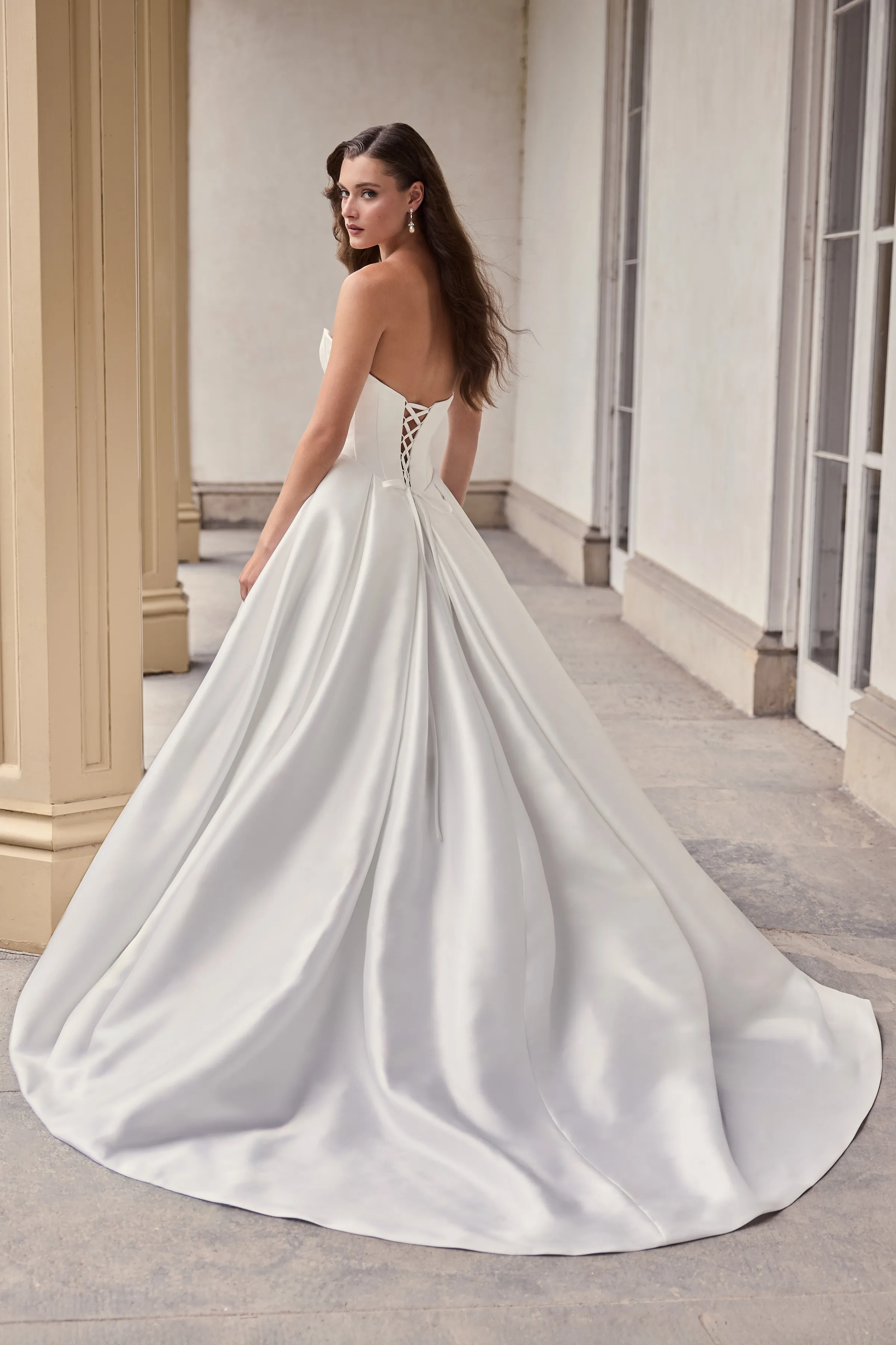 Back view of a strapless ball gown Mikado wedding dress with a lace-up back and a pleated skirt, in front of pillars and a wall with windows.