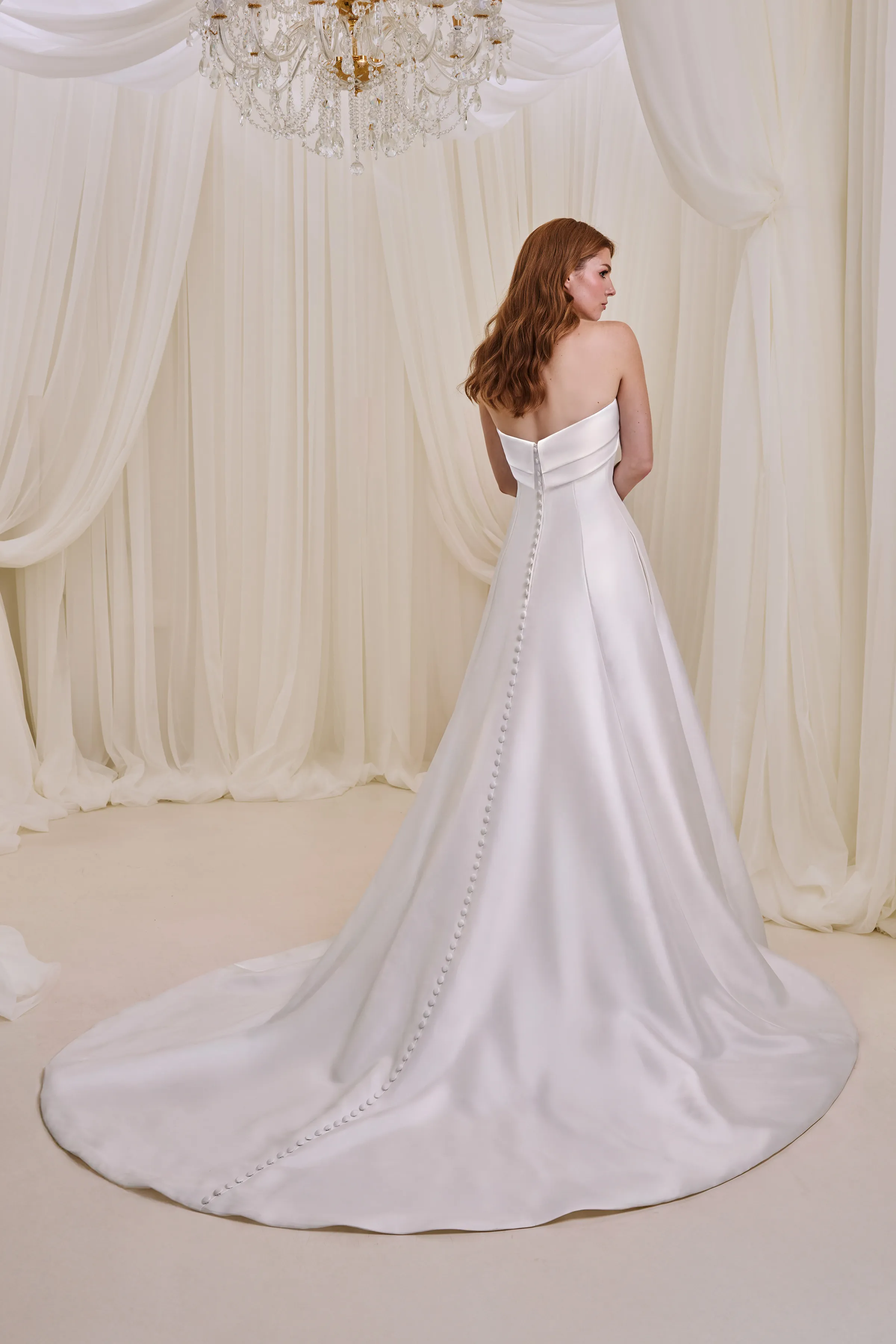 Back view of a strapless Satin wedding dress with buttons down the back of the dress to the hem and an A-line skirt, indoors in front of a wall of tulle curtains.