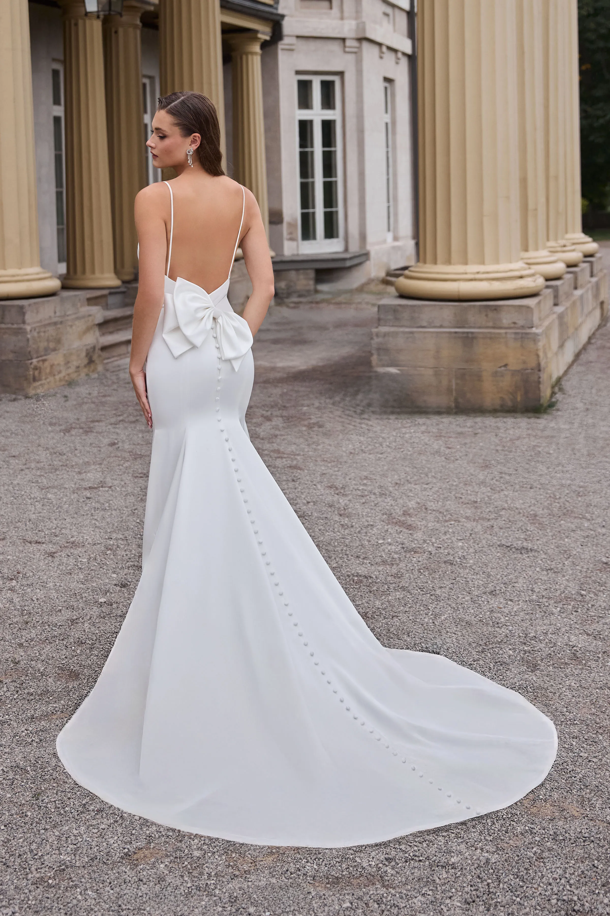 Back view of a Charmeuse wedding dress with an open back, spaghetti straps, and a back bow, with buttons down the skirt of a fitted skirt, outside in front of a building with pillars.