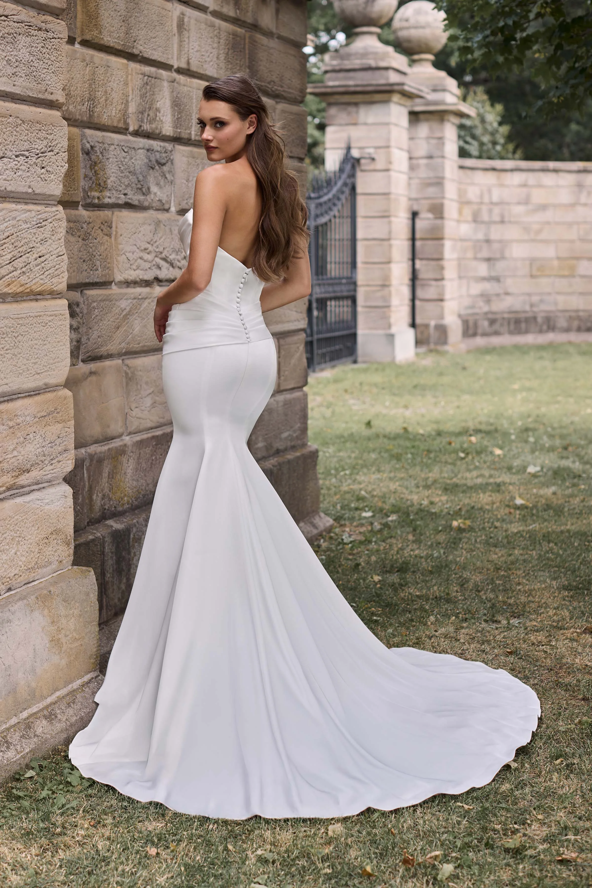 Back view of a strapless Charmeuse wedding dress with buttons down back of bodice and draping at waist, with a fit and flare skirt, outside in front of large stone walls.