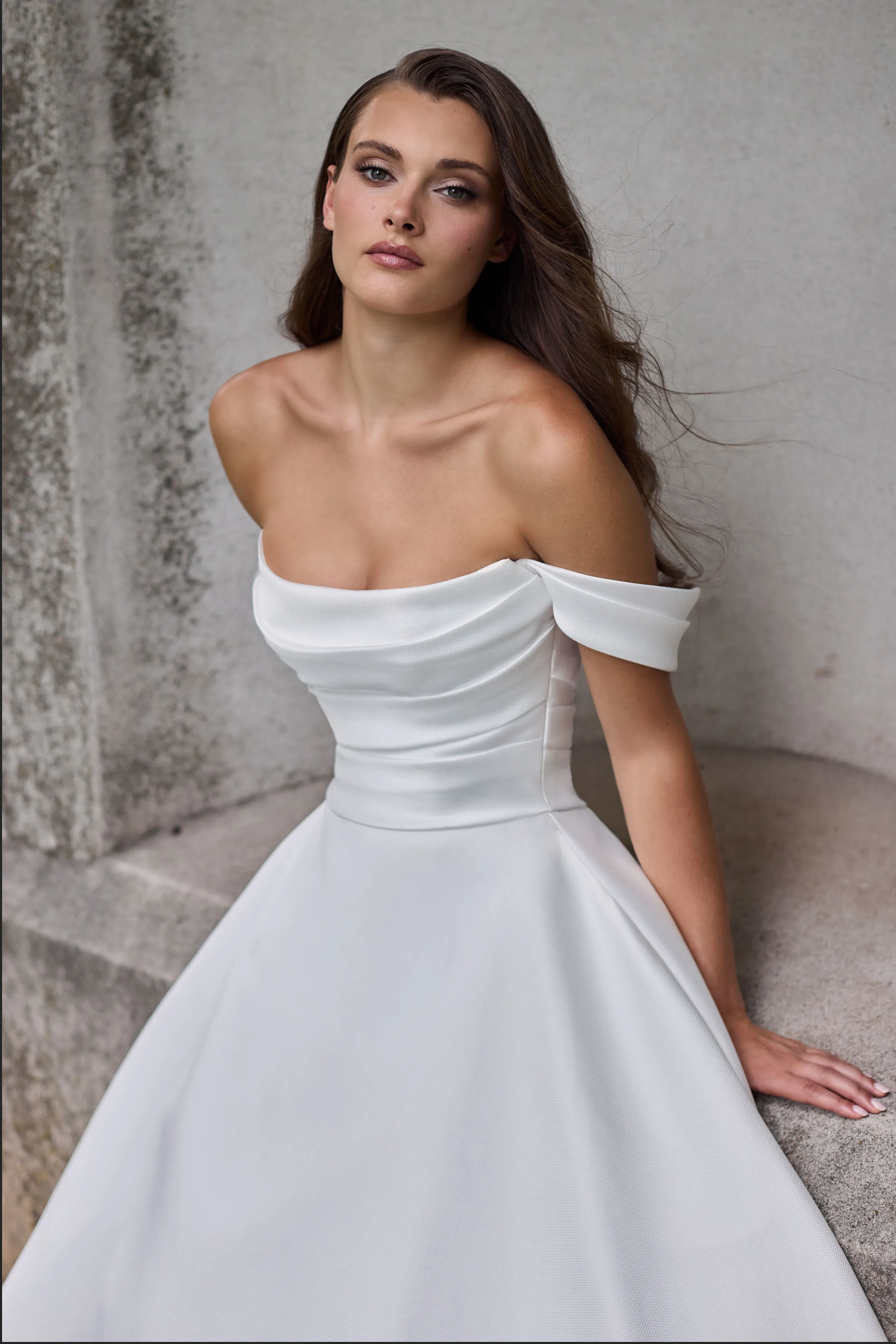 Close up view of a scoop neckline Mikado wedding dress with draped sleeves, a draped bodice, and an A-line skirt, in front of a stone wall.