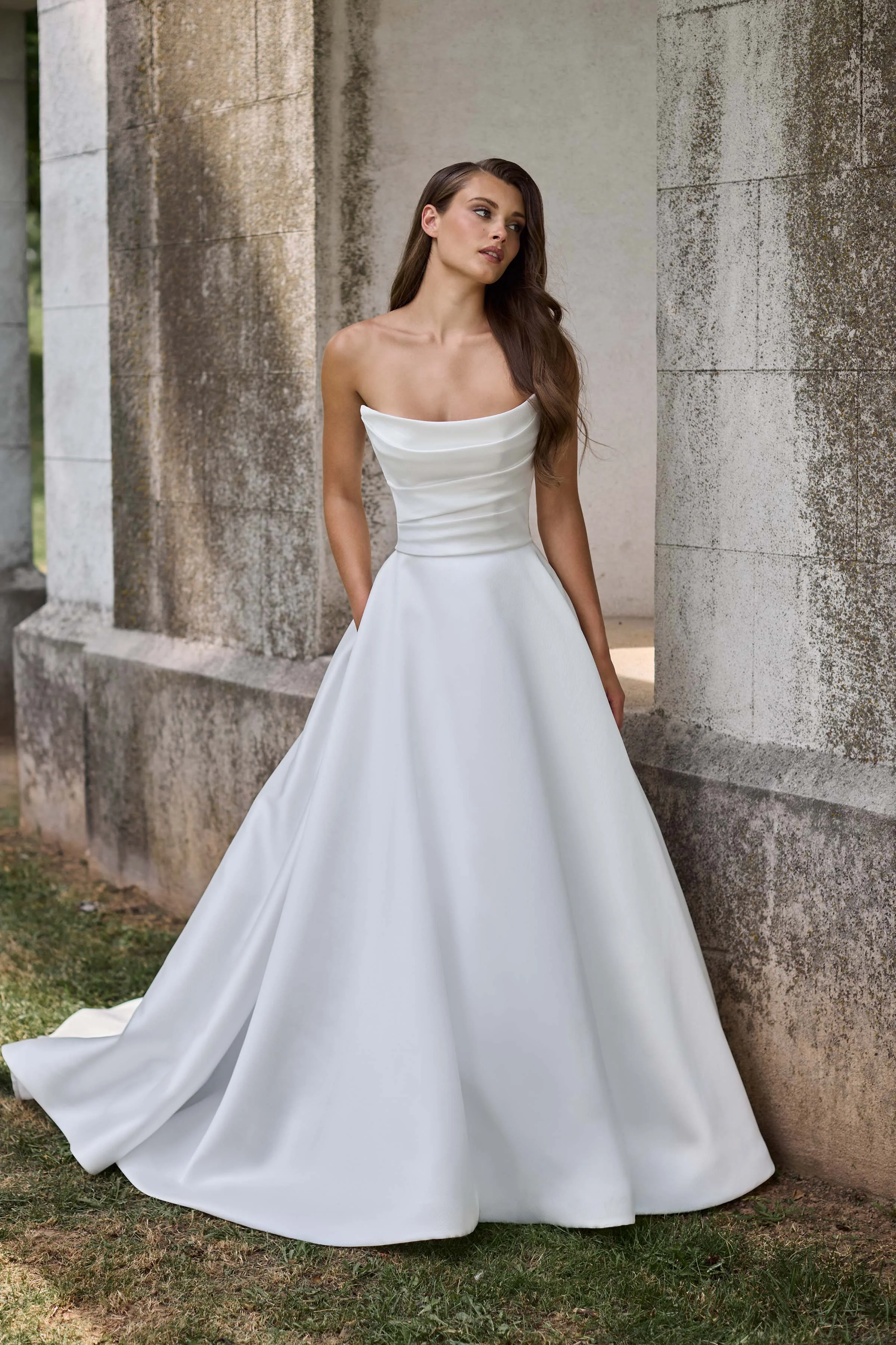 Front view of a strapless scoop neckline Mikado wedding dress with a draped bodice and an A-line skirt with pockets, outside in front of a stone wall.