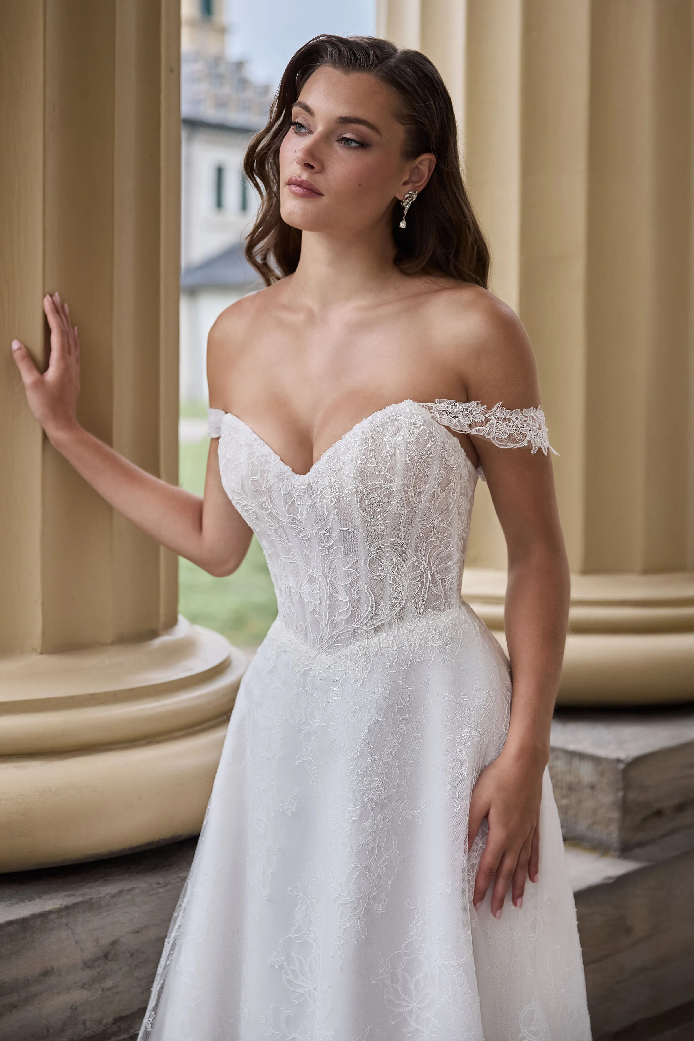 Close up view of a strapless Lace wedding dress with draped sleeves, a Basque waistline, and a Lace A-line skirt, outside in front of pillars with a manor in the background.