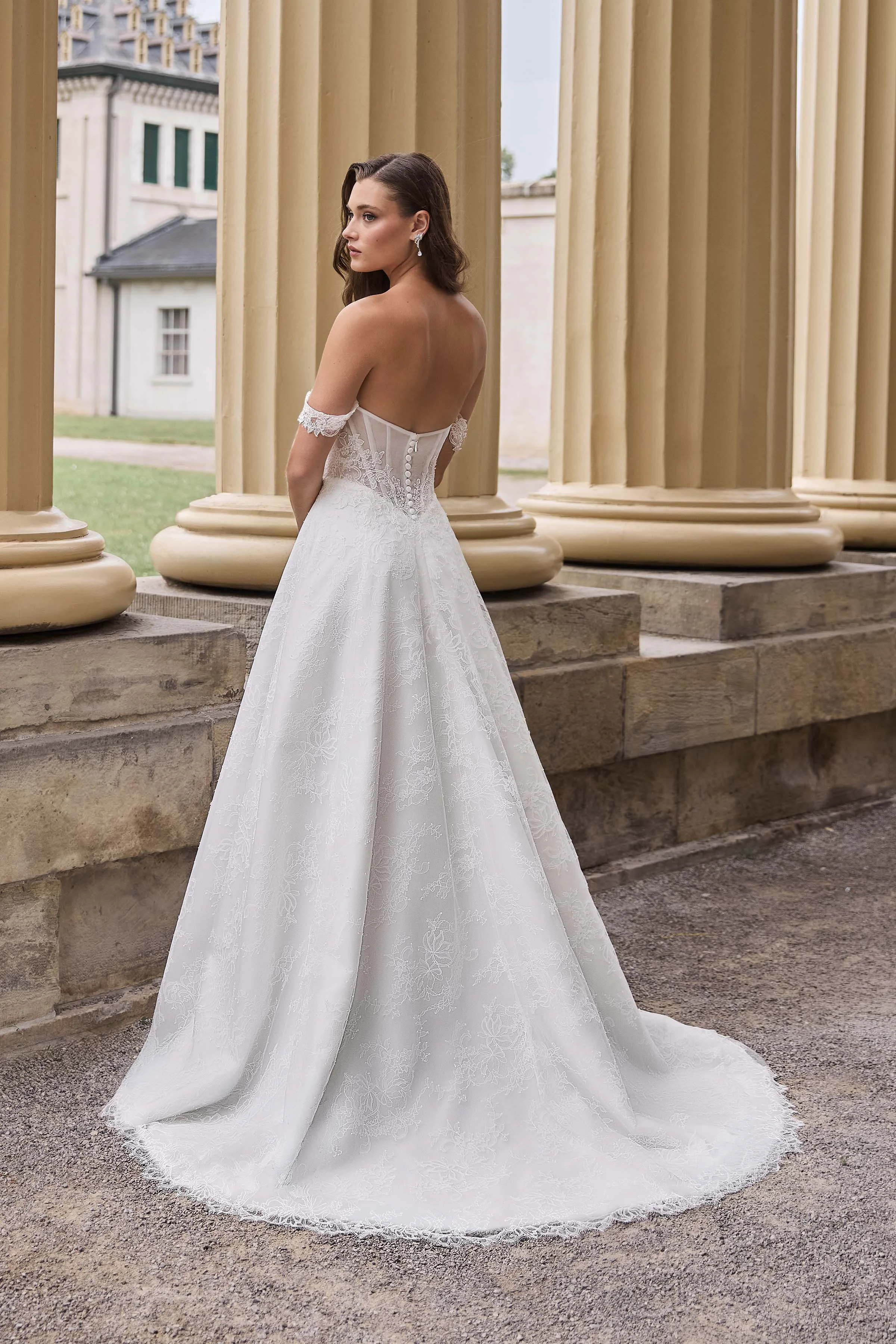 Back view of a strapless Lace wedding dress with draped sleeves, a sheer lace back with buttons on the back of the bodice, and a Lace A-line skirt, outside in front of pillars with a manor in the background.