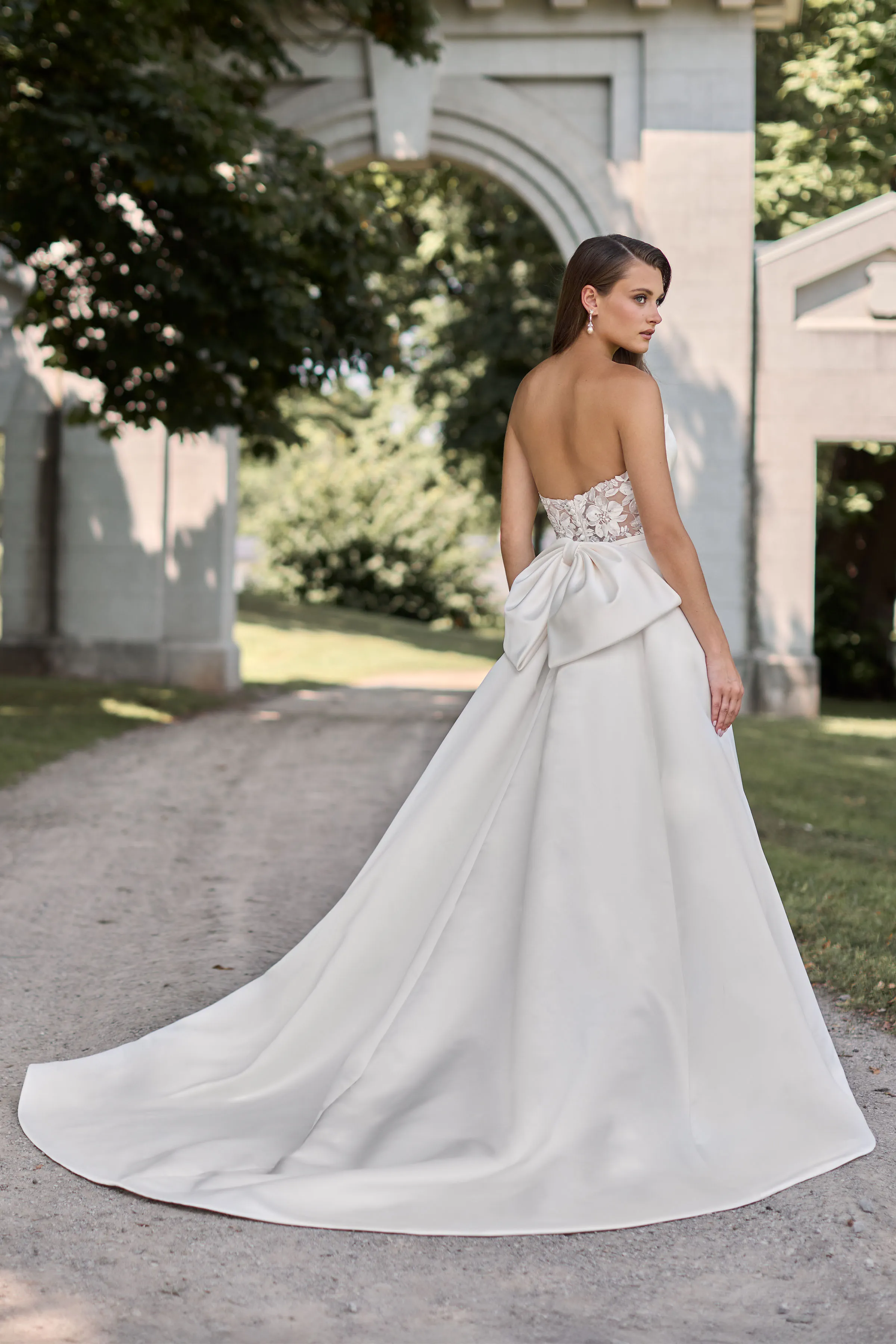 Back view of a strapless Duchesse Satin wedding dress with a sheer lace back, a large back bow, and an A-line skirt, outside on a path in front of a stone arch.