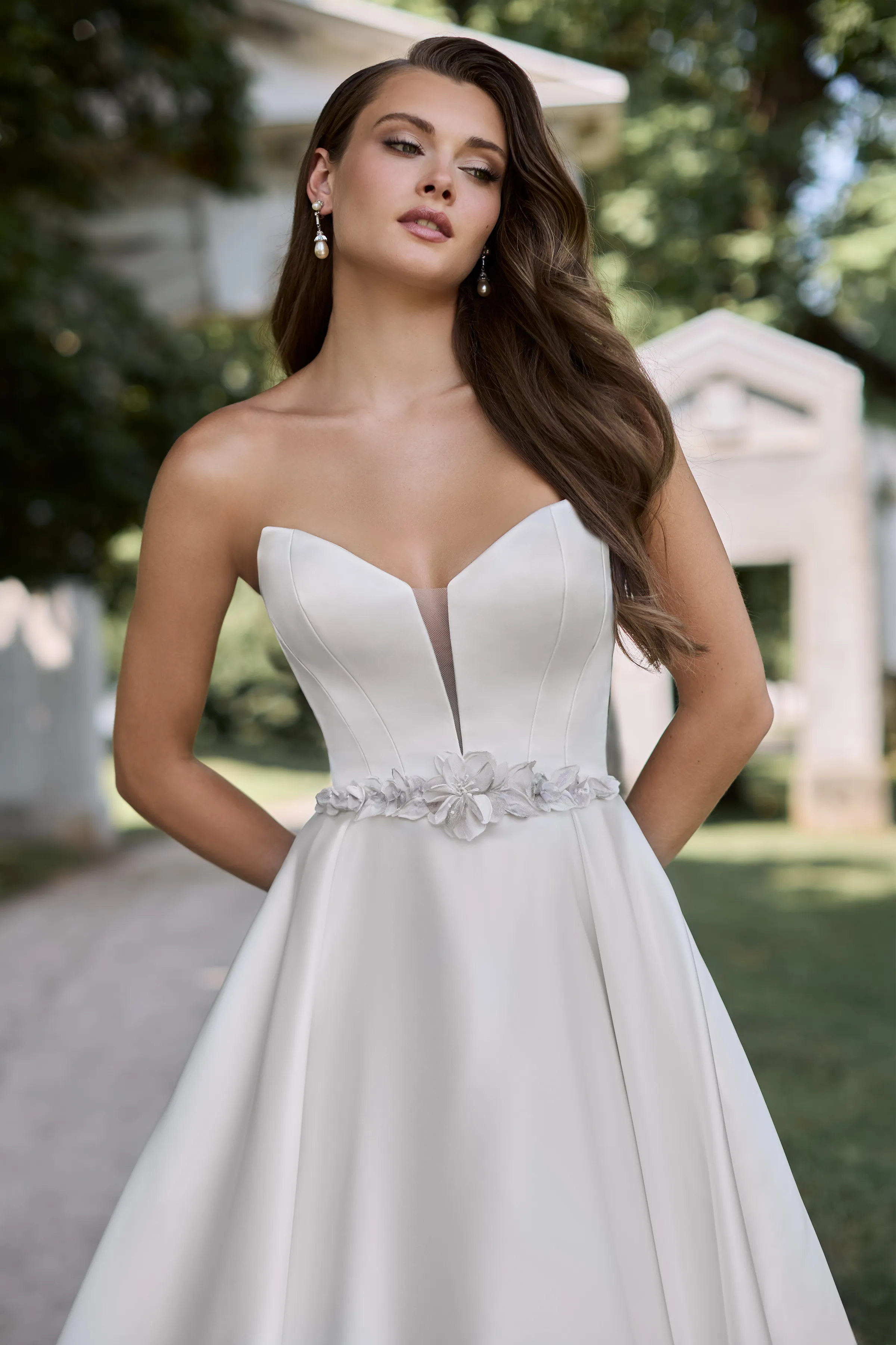 Close up view of a strapless Duchesse Satin wedding dress with a plunging neckline, a floral applique belt, and a pleated skirt with pockets, outside on a path in front of a stone arch.