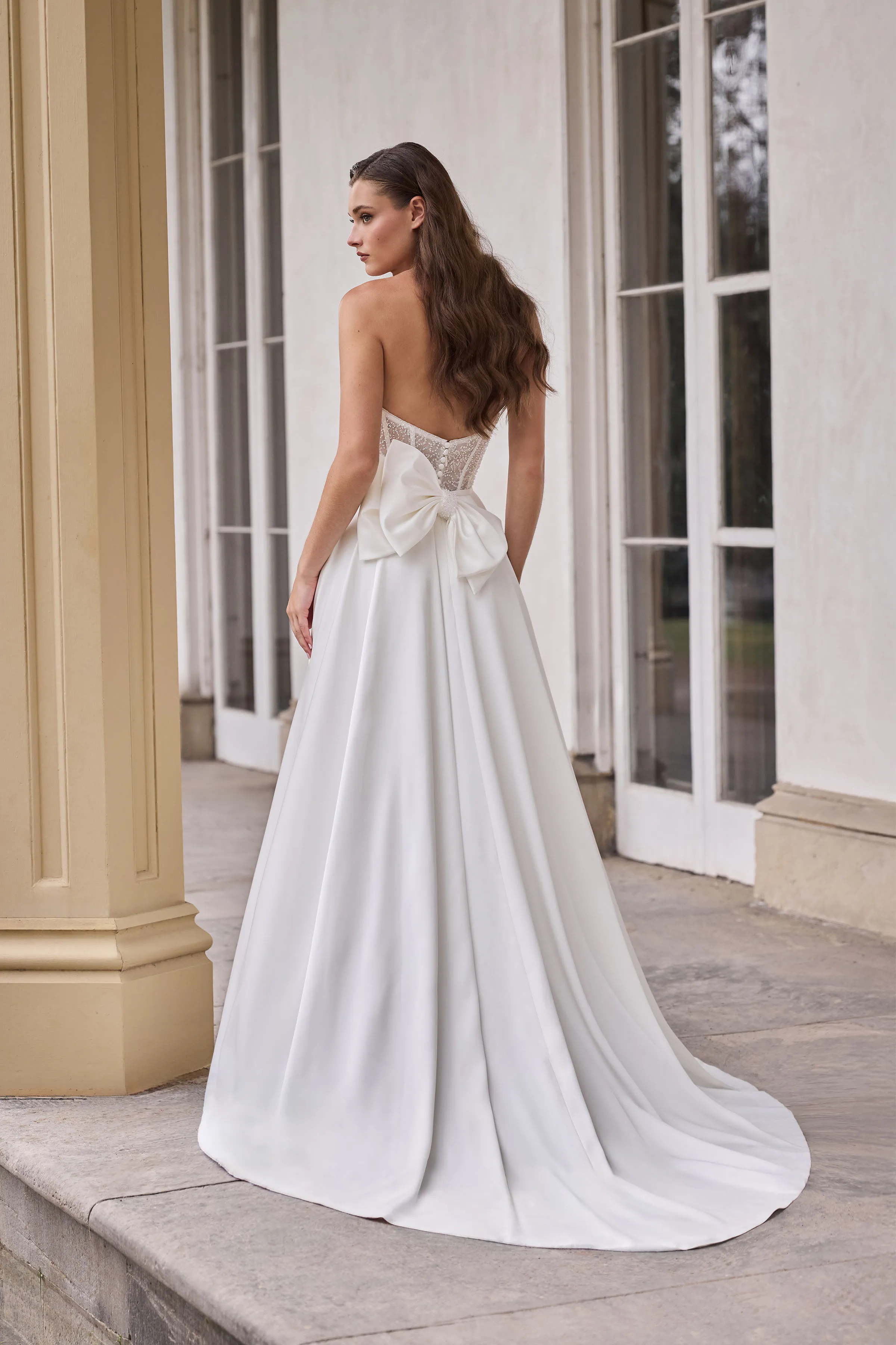 Back view of a strapless wedding dress with a sheer Beaded Tulle corset bodice with buttons, a back bow, and an A-line skirt, in front of pillars and a wall with windows.