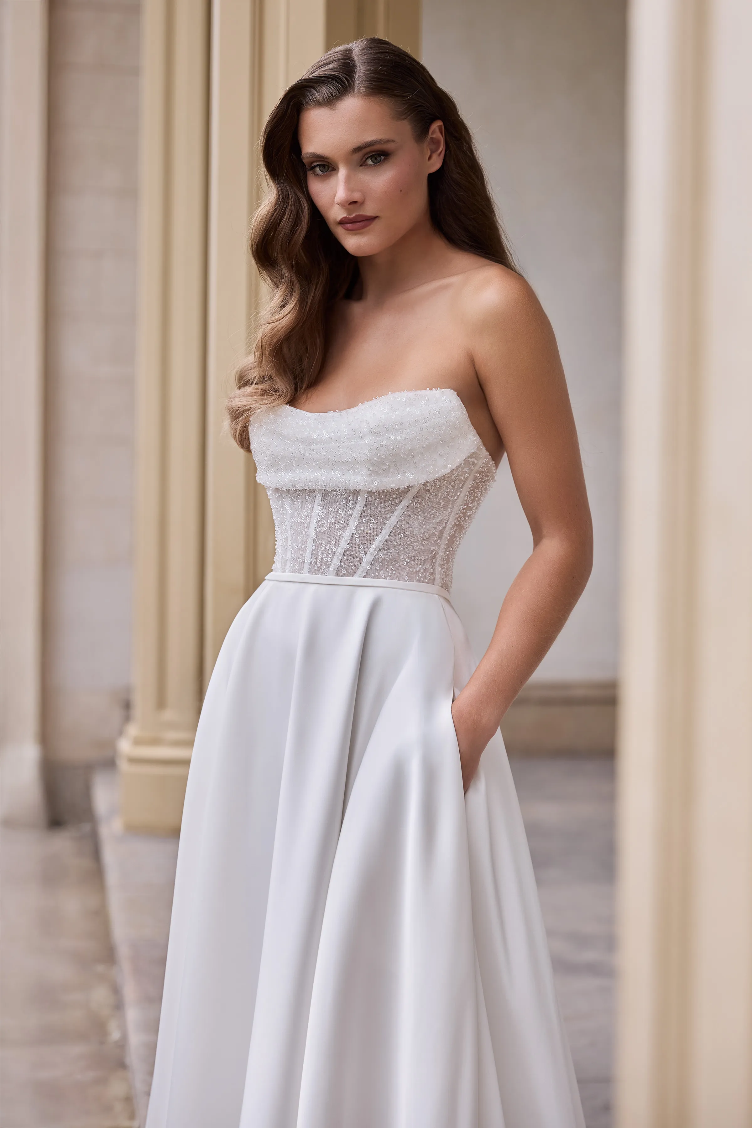 Close up view of a strapless wedding dress with a sheer Beaded Tulle corset bodice, a fabric band at the waist, and a Charmeuse A-line skirt with pockets, in front of the outside of a pillared building.