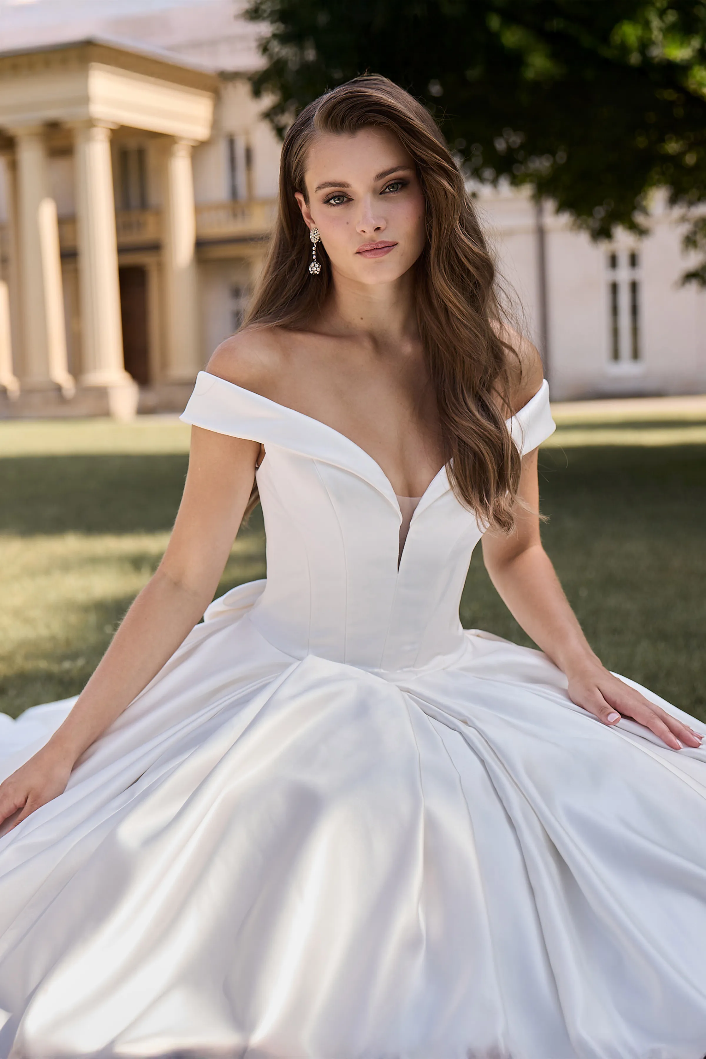 Close up view of a model sitting on a grass lawn wearing an off the shoulder Satin wedding dress with a plunging neckline and seamed bodice, and a pleated skirt, in front of a manor.