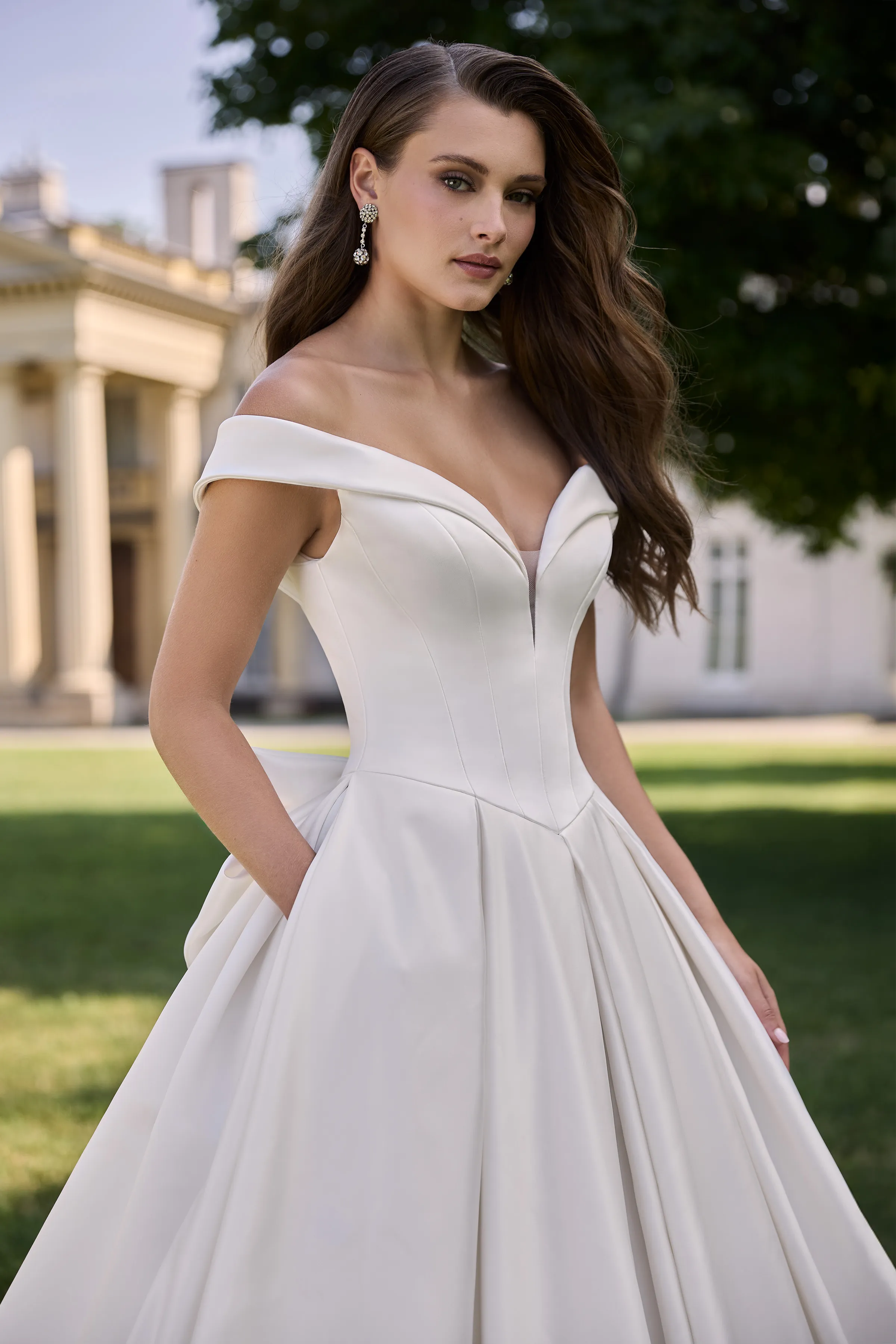 Close up view of an off the shoulder Satin wedding dress with a plunging neckline, seamed bodice, Basque waistline, and a pleated ball gown skirt with pockets, outside in front of a manor.
