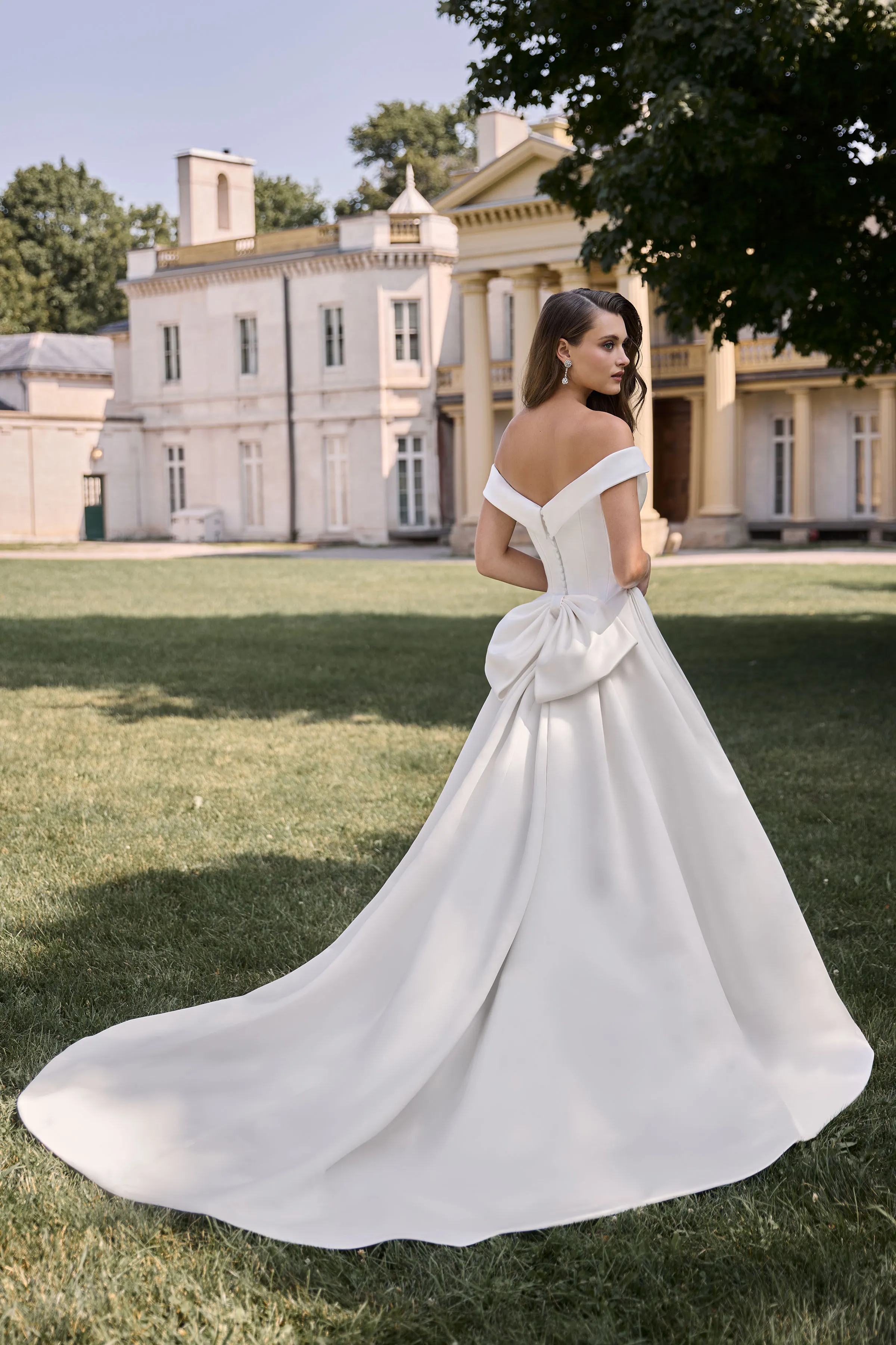 Back view of an off the shoulder Satin wedding dress with buttons down the back of the bodice, a back bow, and a ball gown skirt, outside in front of a manor.