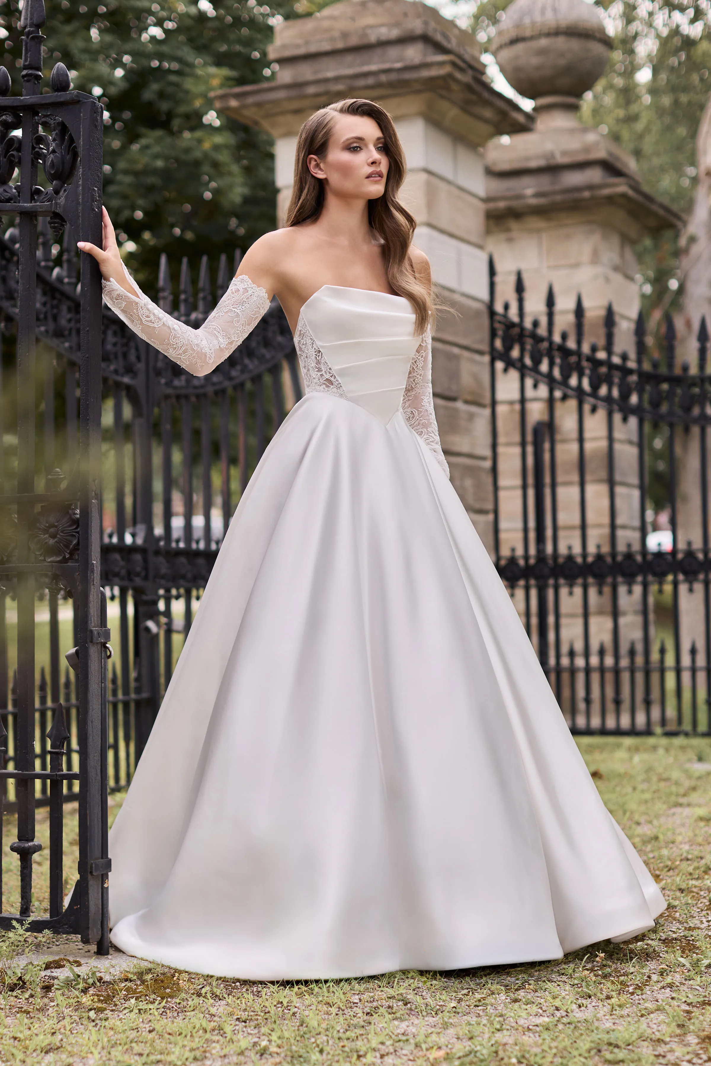 Front view of a strapless Satin wedding dress with lace cutouts on the draped bodice, separate lace sleeves, and a full A-line skirt, outside in front of iron gates.