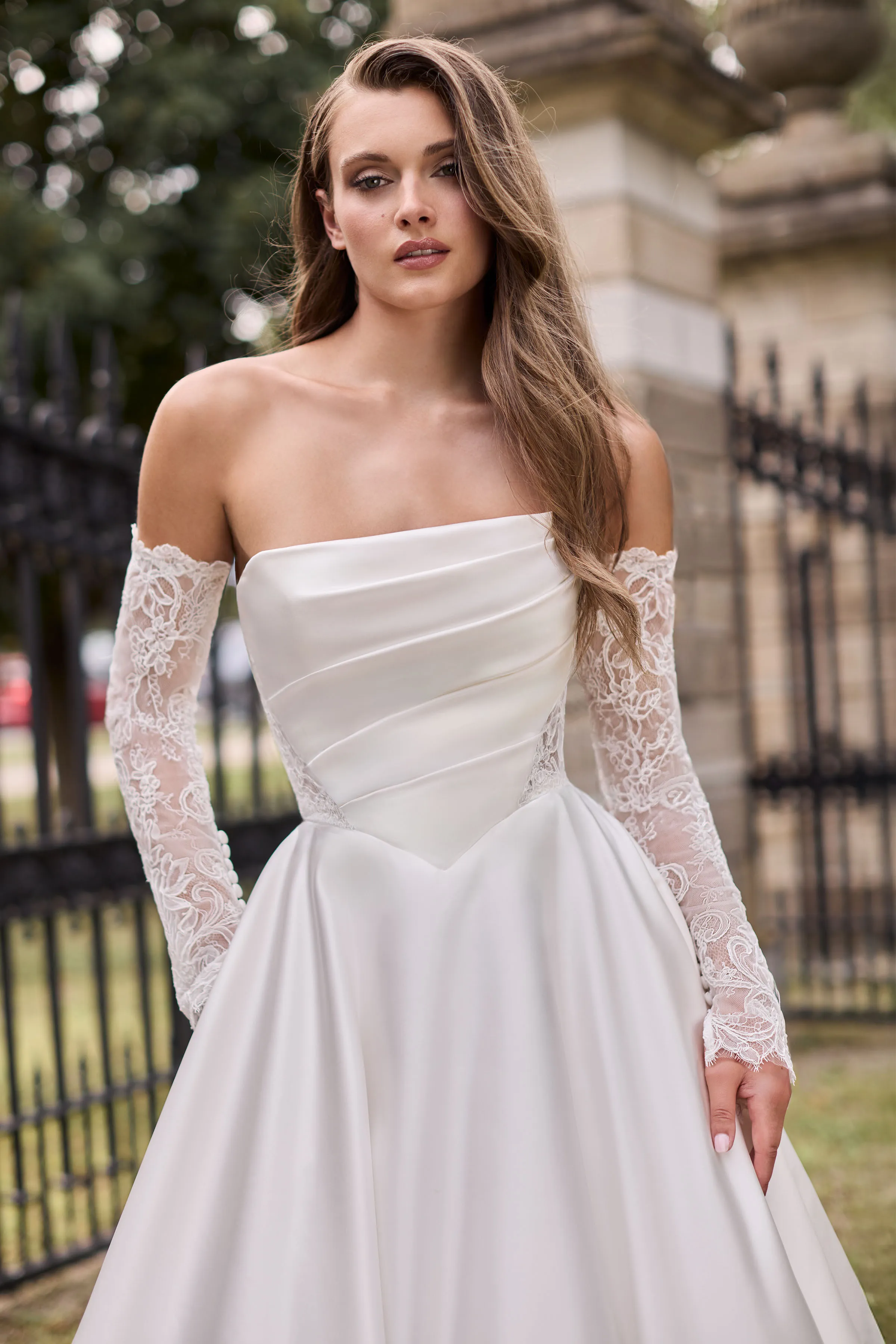Close up view of a strapless Satin wedding dress with a straight neckline, draped bodice, and lace cutouts on the bodice, with separate lace sleeves and an A-line skirt, outside in front of iron gates.