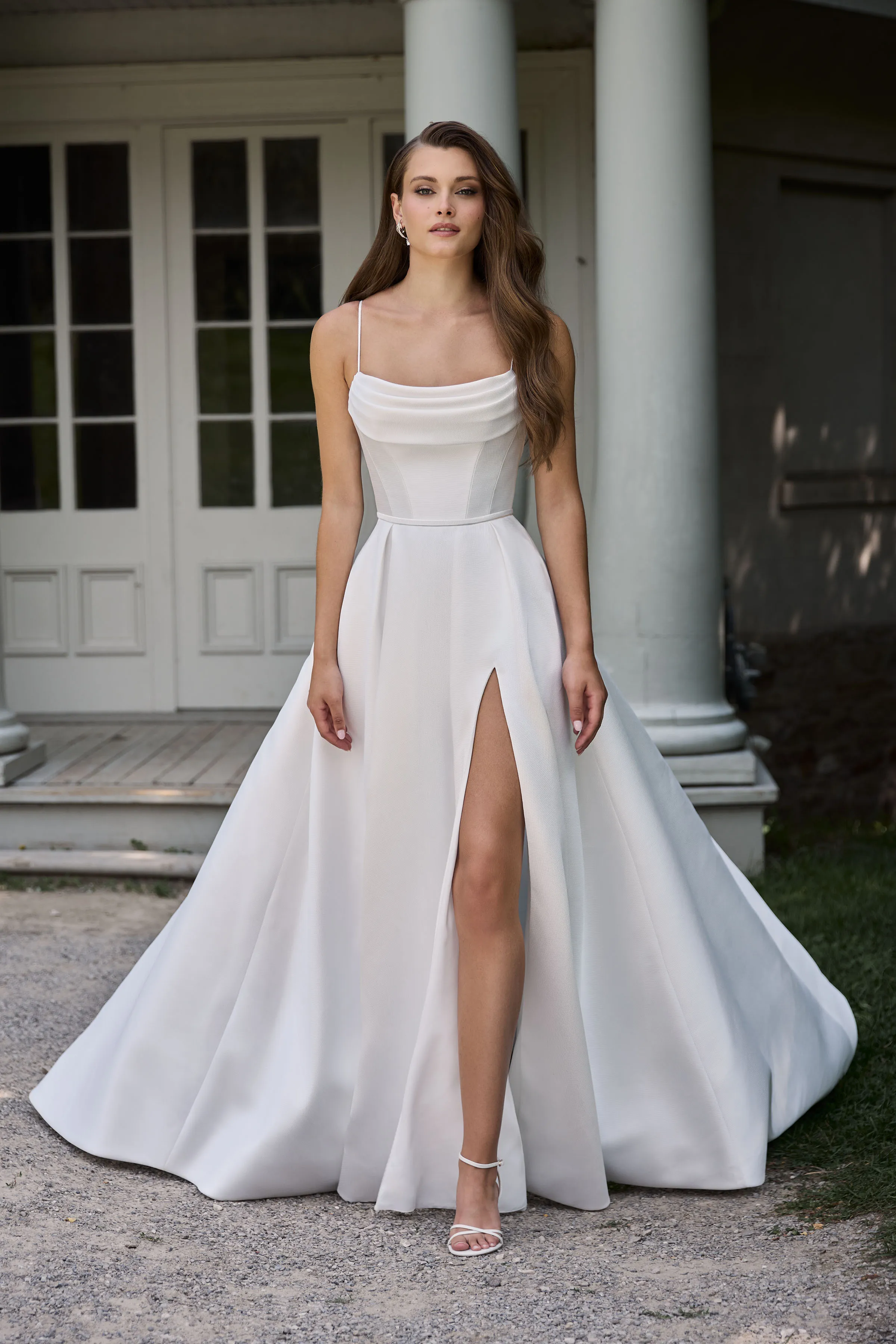 Front view of a Mikado wedding dress with spaghetti straps, draped neckline, and a full skirt with a skirt slit, in front of a building with pillars.