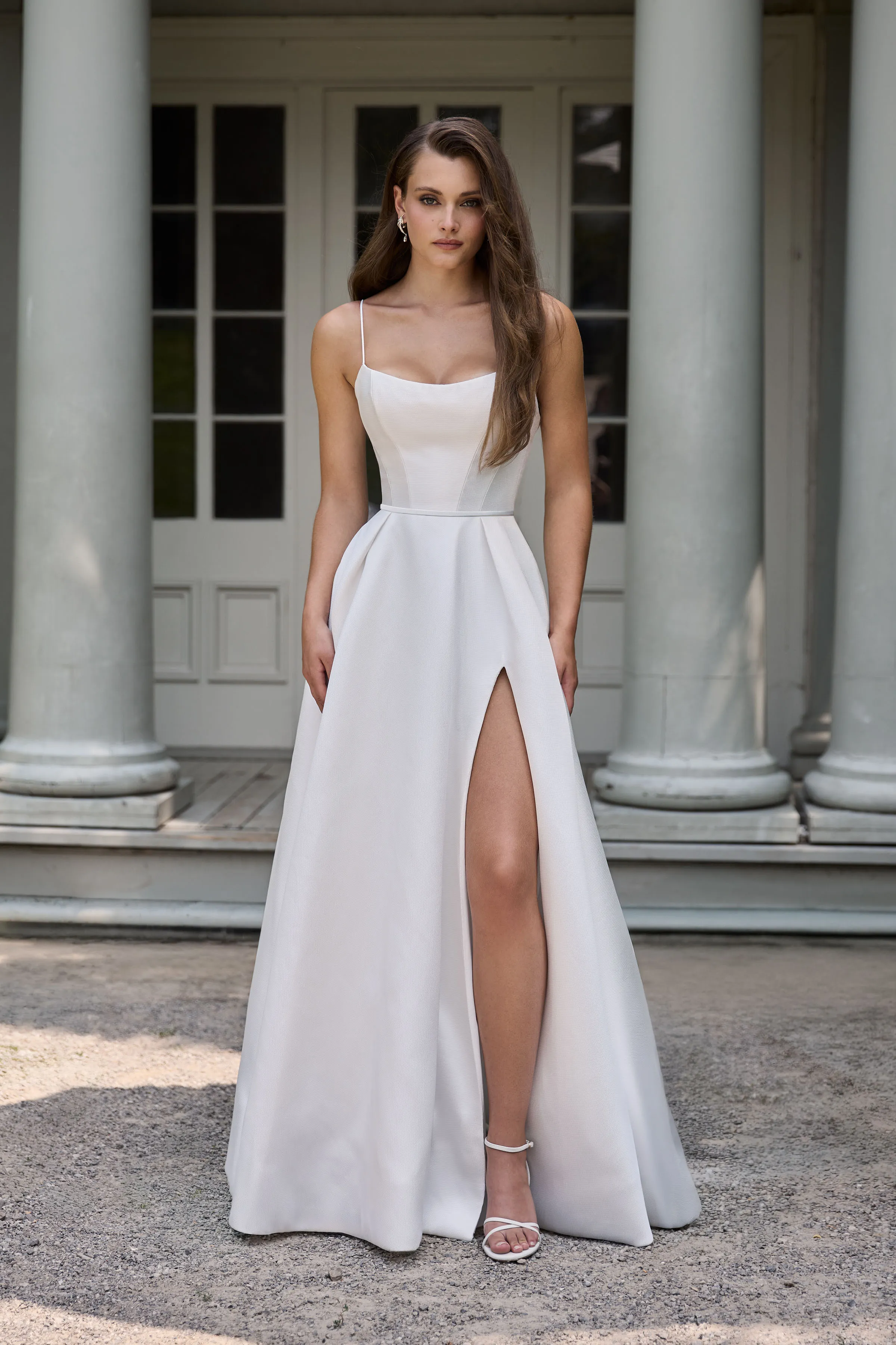 Front view of a Mikado wedding dress with spaghetti straps, draped neckline, and a full skirt with a skirt slit, in front of a building with pillars.