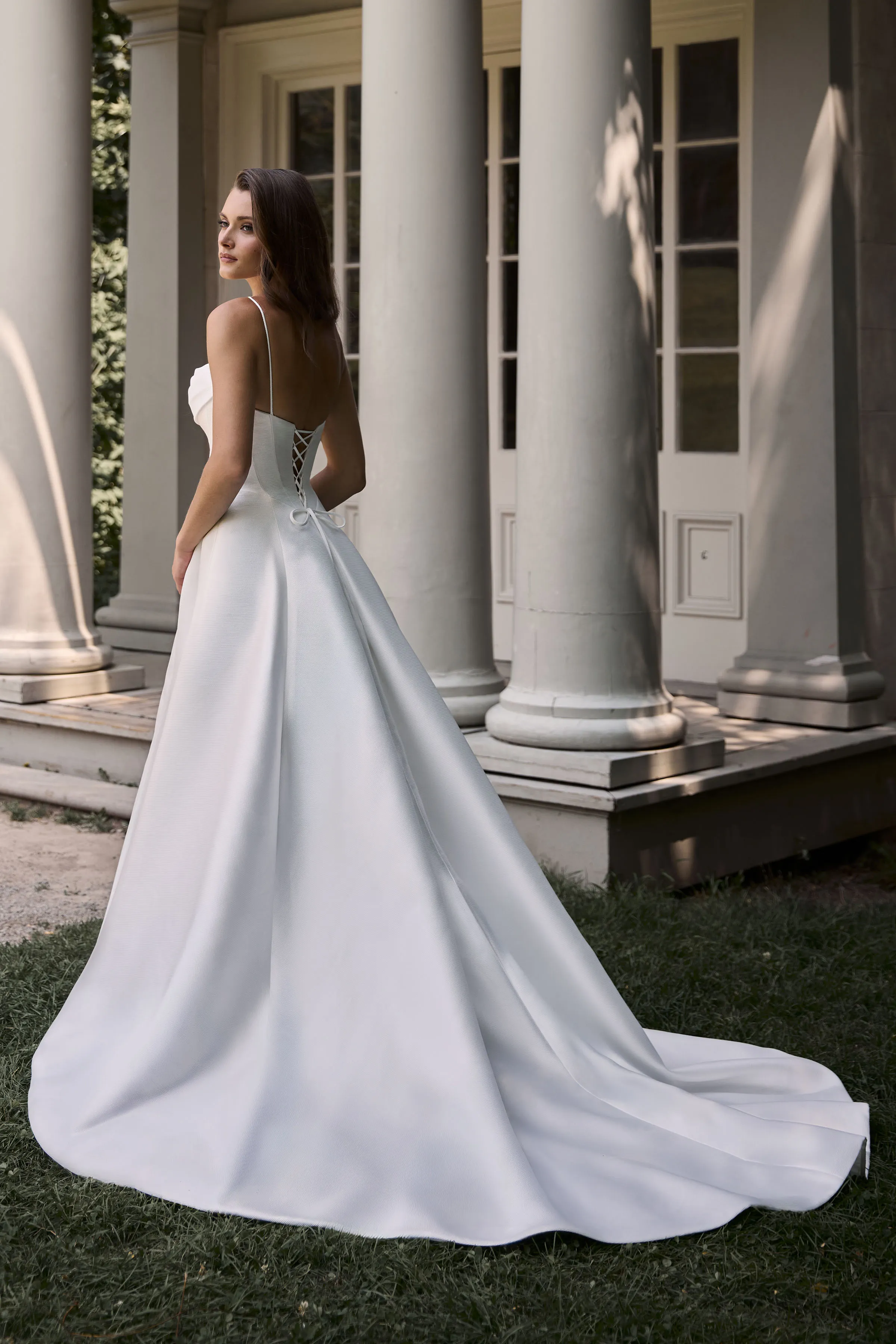 Back view of a Mikado wedding dress with spaghetti straps, a lace-up back, and a full skirt with pleats, in front of a building with pillars.