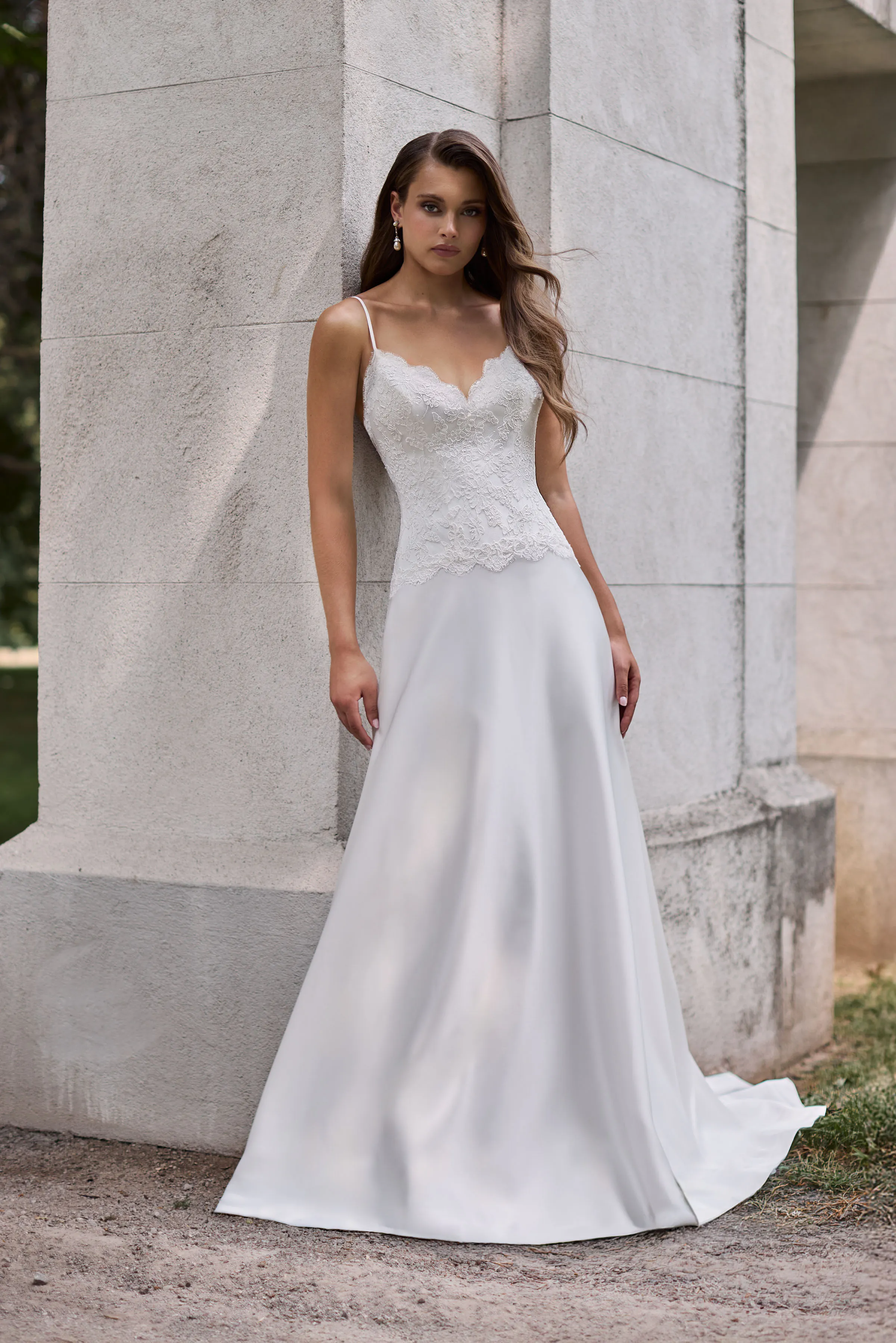 Front view of a Lace bodice wedding dress with spaghetti straps, scallop along the neckline and bottom of the bodice, and a full Charmeuse circle skirt, outside in front of a large stone pillar.