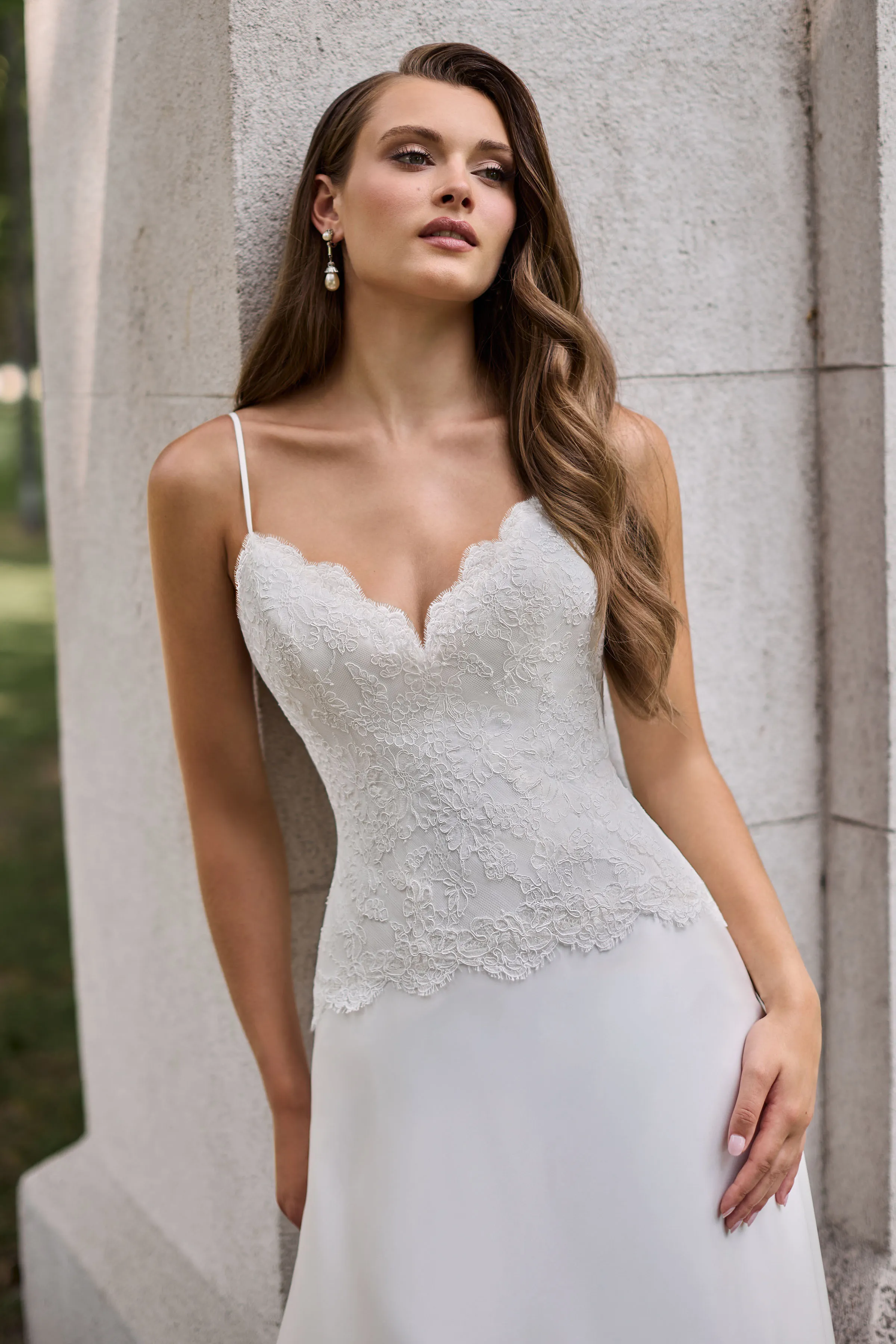 Close up view of a Lace bodice wedding dress with spaghetti straps, scallop along the neckline and bottom of the bodice, and a Charmeuse skirt, outside in front of a large stone pillar.
