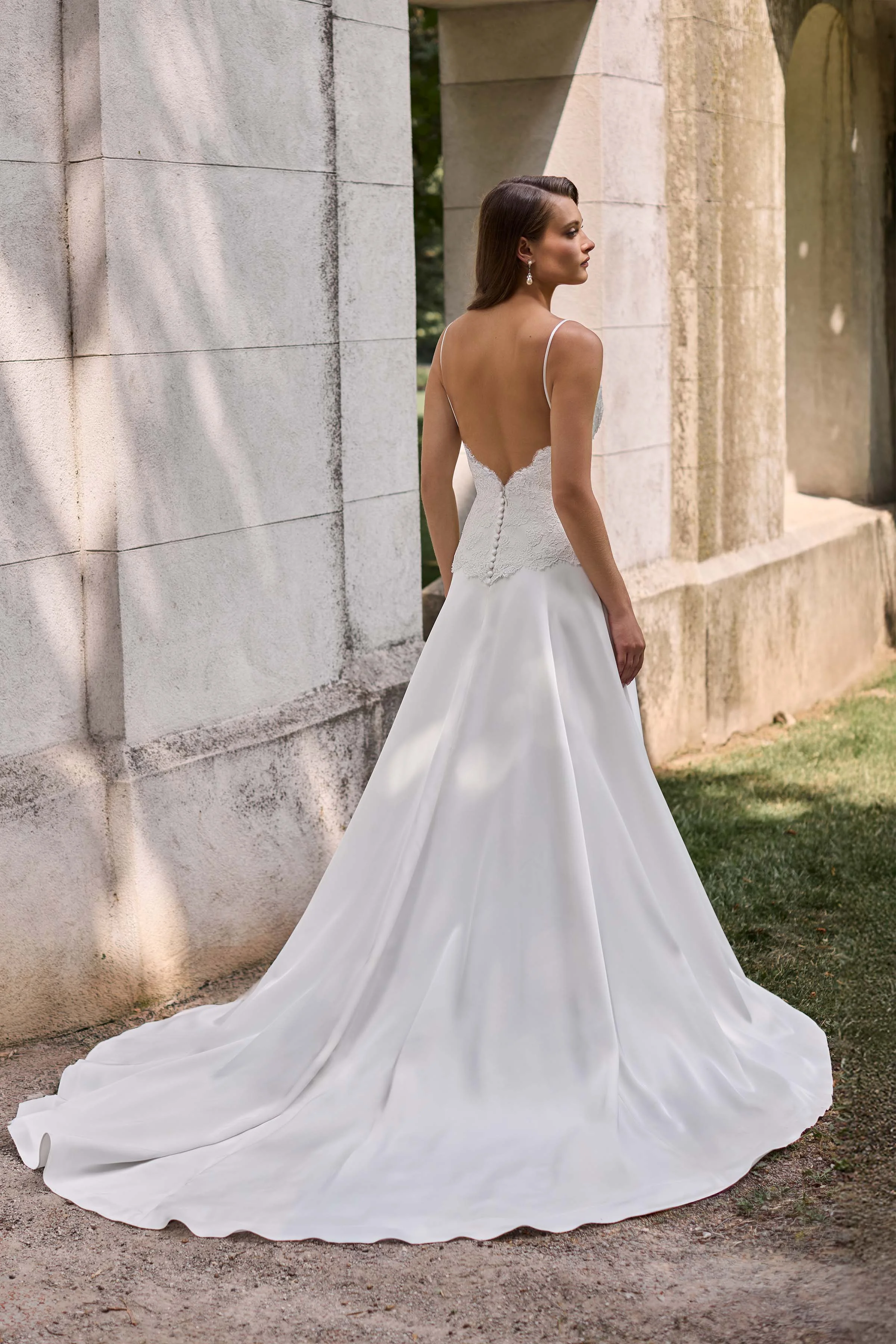Back view of a Lace bodice wedding dress with spaghetti straps and buttons down the back of the bodice, with a full Charmeuse circle skirt, outside in front a large stone wall.