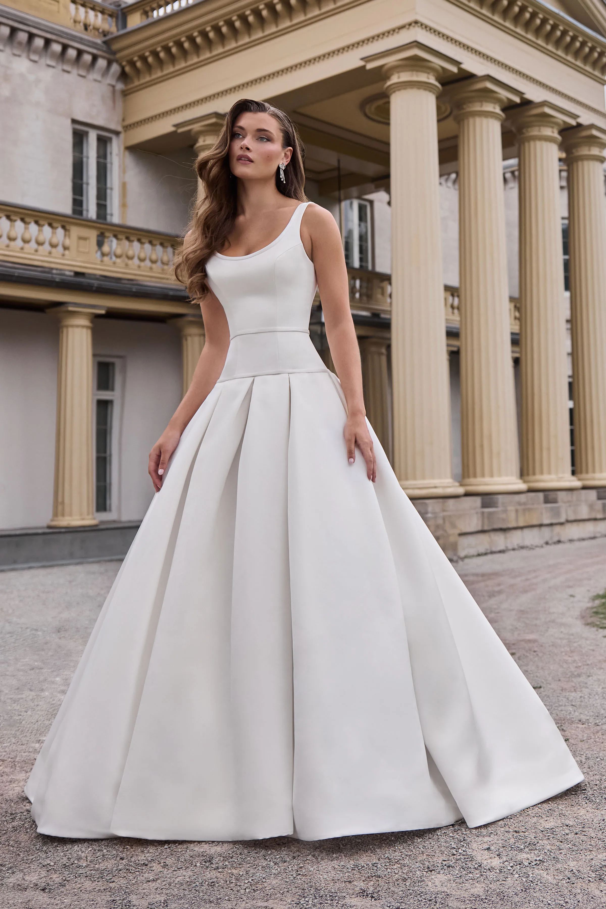 Front view of a sleeveless Satin wedding dress with a scoop neckline, a drop waisted bodice, and a pleated skirt, outside in front of a building with pillars.