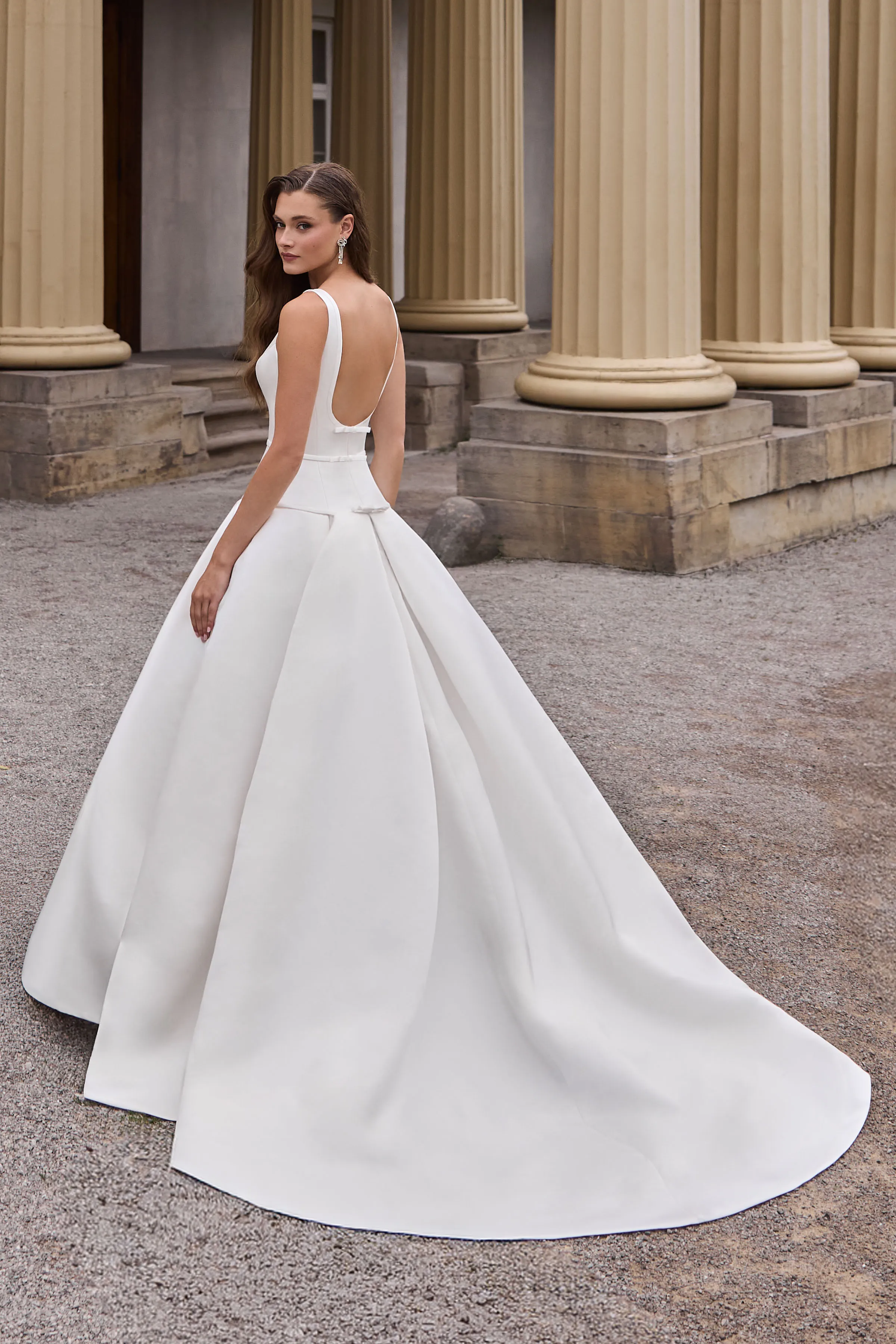 Back view of a sleeveless Satin wedding dress with a drop waisted bodice, with three small bows at the back and a pleated skirt, outside in front of a building with pillars.
