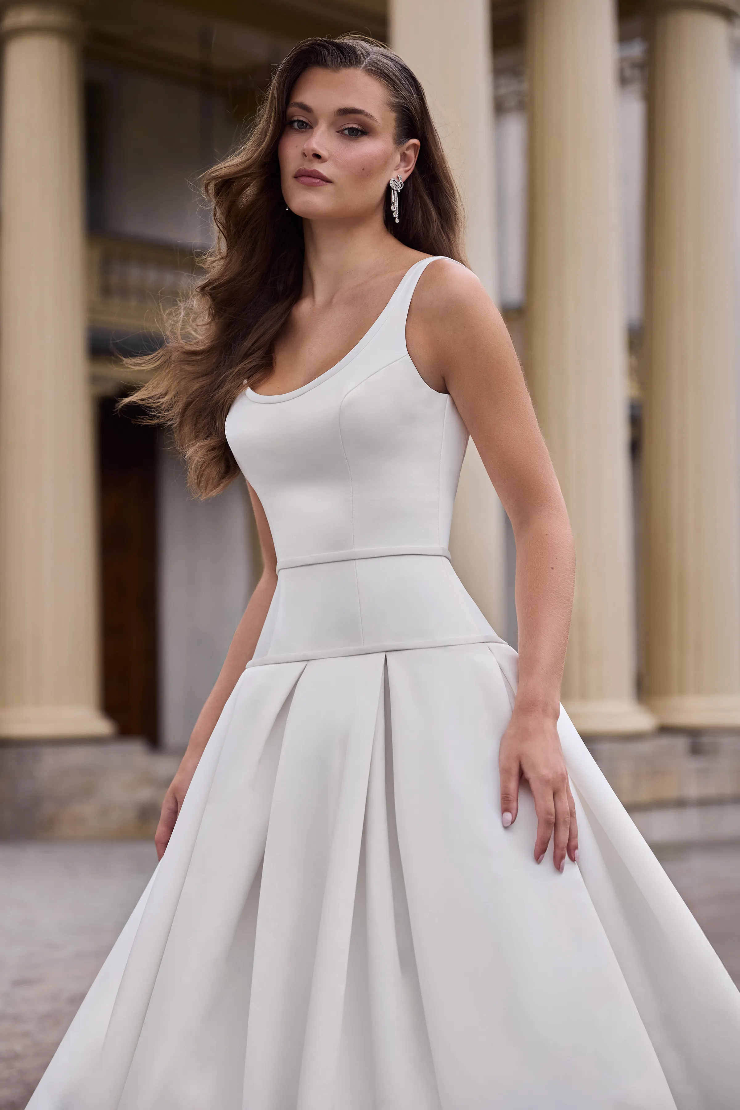Close up view of a sleeveless Satin wedding dress with a scoop neckline, a drop waisted bodice, and a pleated skirt, outside in front of a building with pillars.