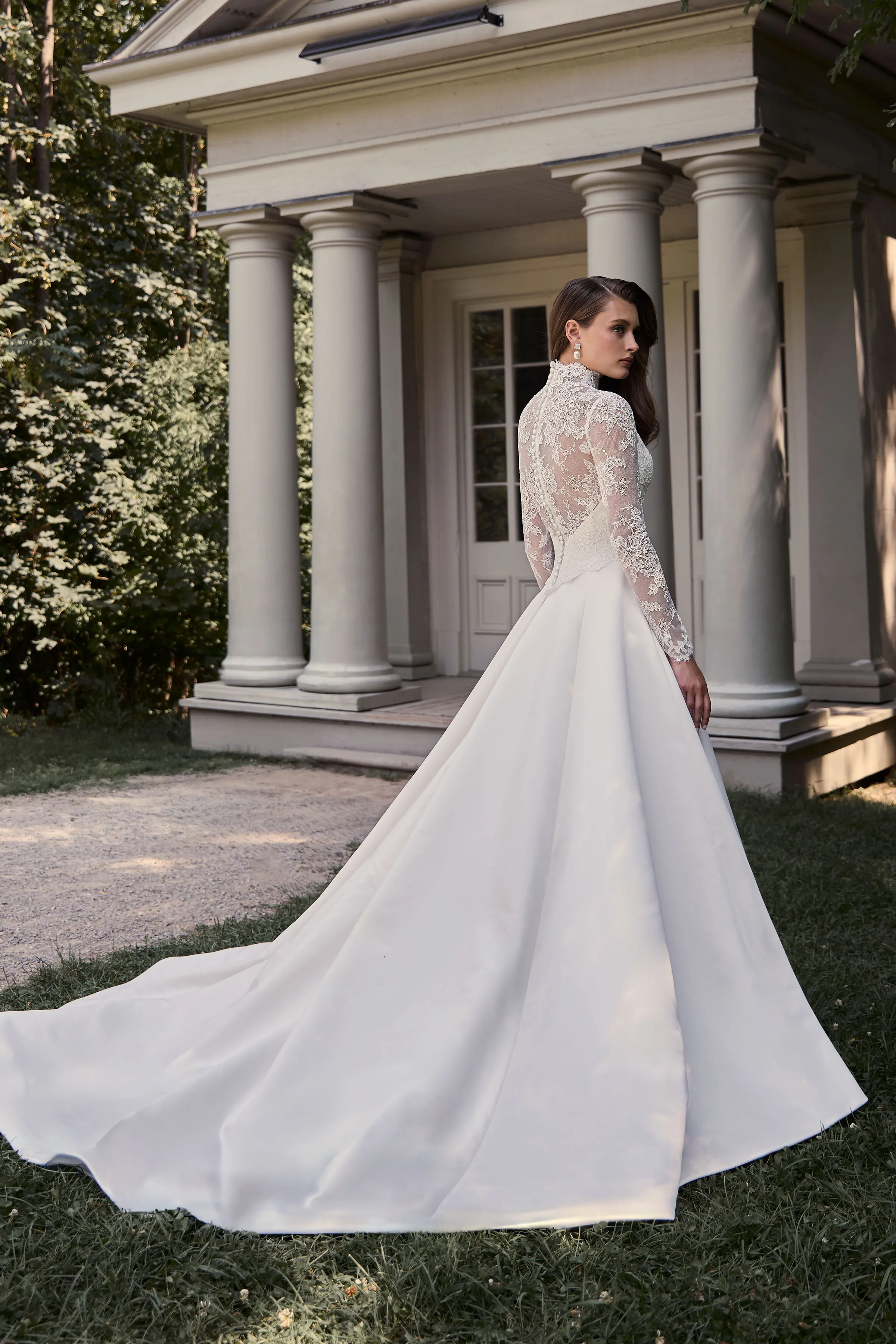 Back view of a Satin wedding dress with long lace sleeves, a high neckline, and sheer lace back with buttons down the bodice, and a Satin A-line skirt, outside in front of a building with pillars.