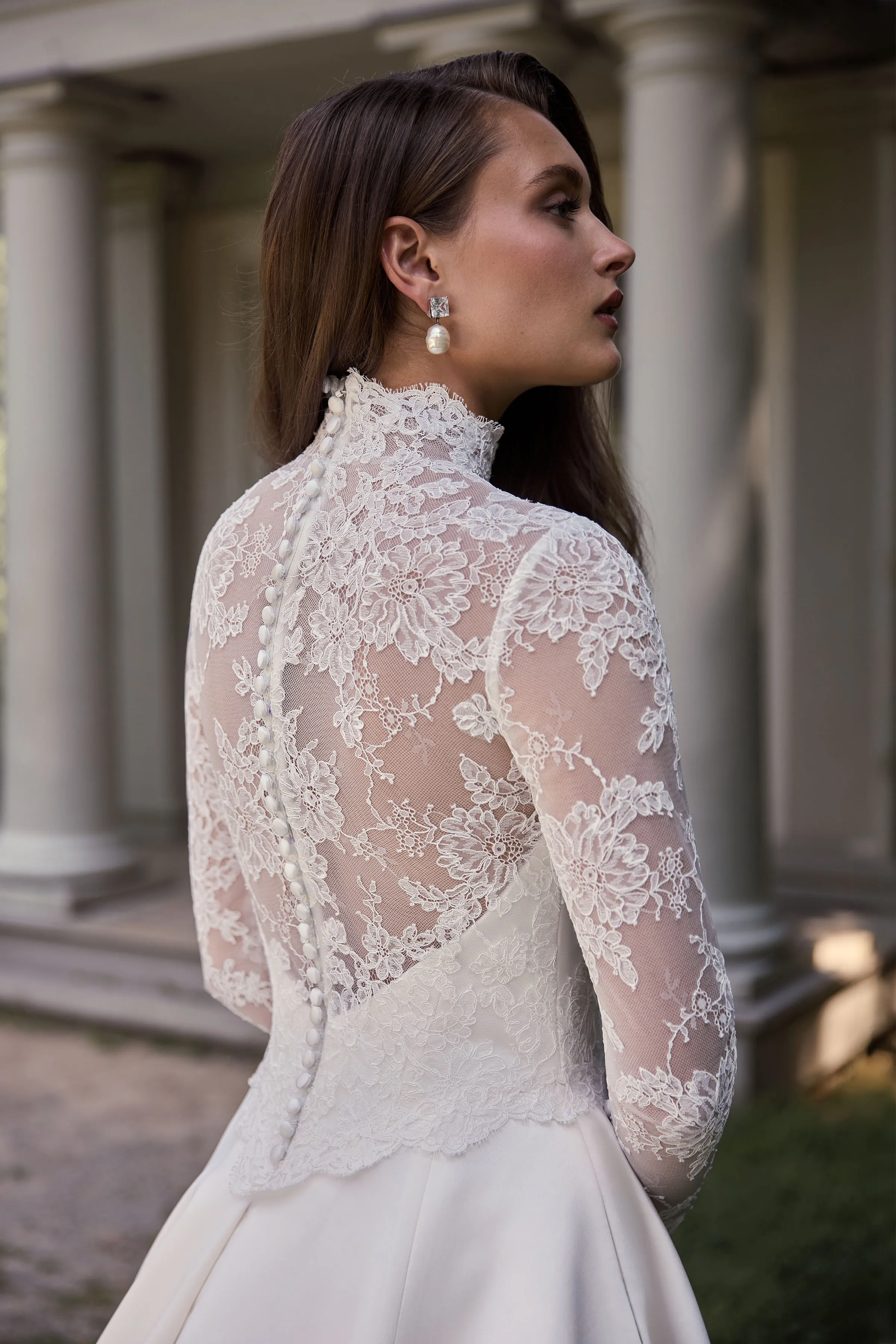 Close up back view of a Satin wedding dress with long lace sleeves, a high neckline, and sheer lace back with buttons down the bodice, and a Satin skirt, outside in front of a building with pillars.