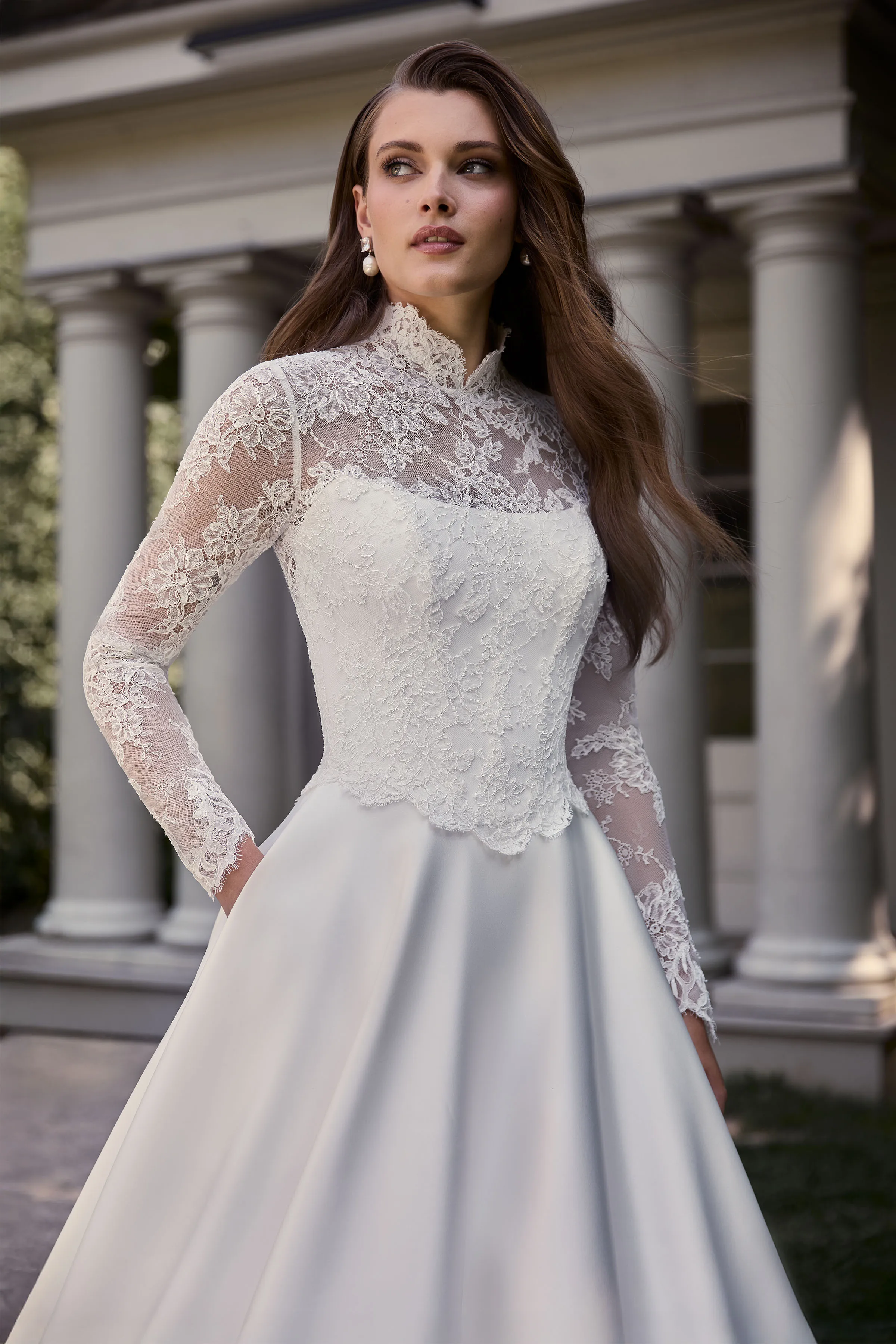 Close up view of a Satin wedding dress with a long sleeve lace bodice, high neckline, and Satin A-line skirt with pockets, outside in front of a building with pillars.