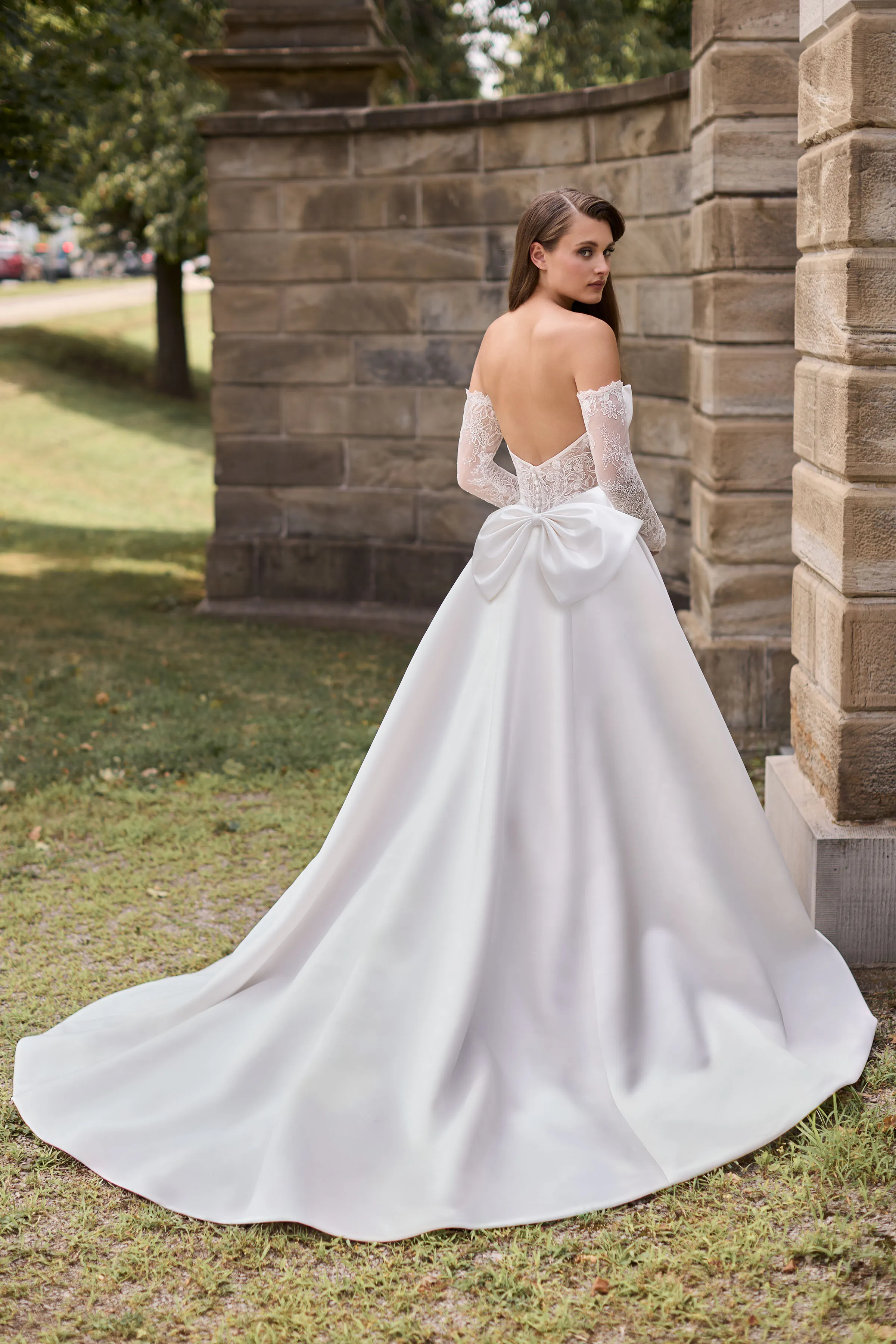 Back view of a strapless Satin wedding dress with a sheer lace back, buttons on the back of the bodice, a back bow, and an A-line skirt, outside in front of a stone wall.